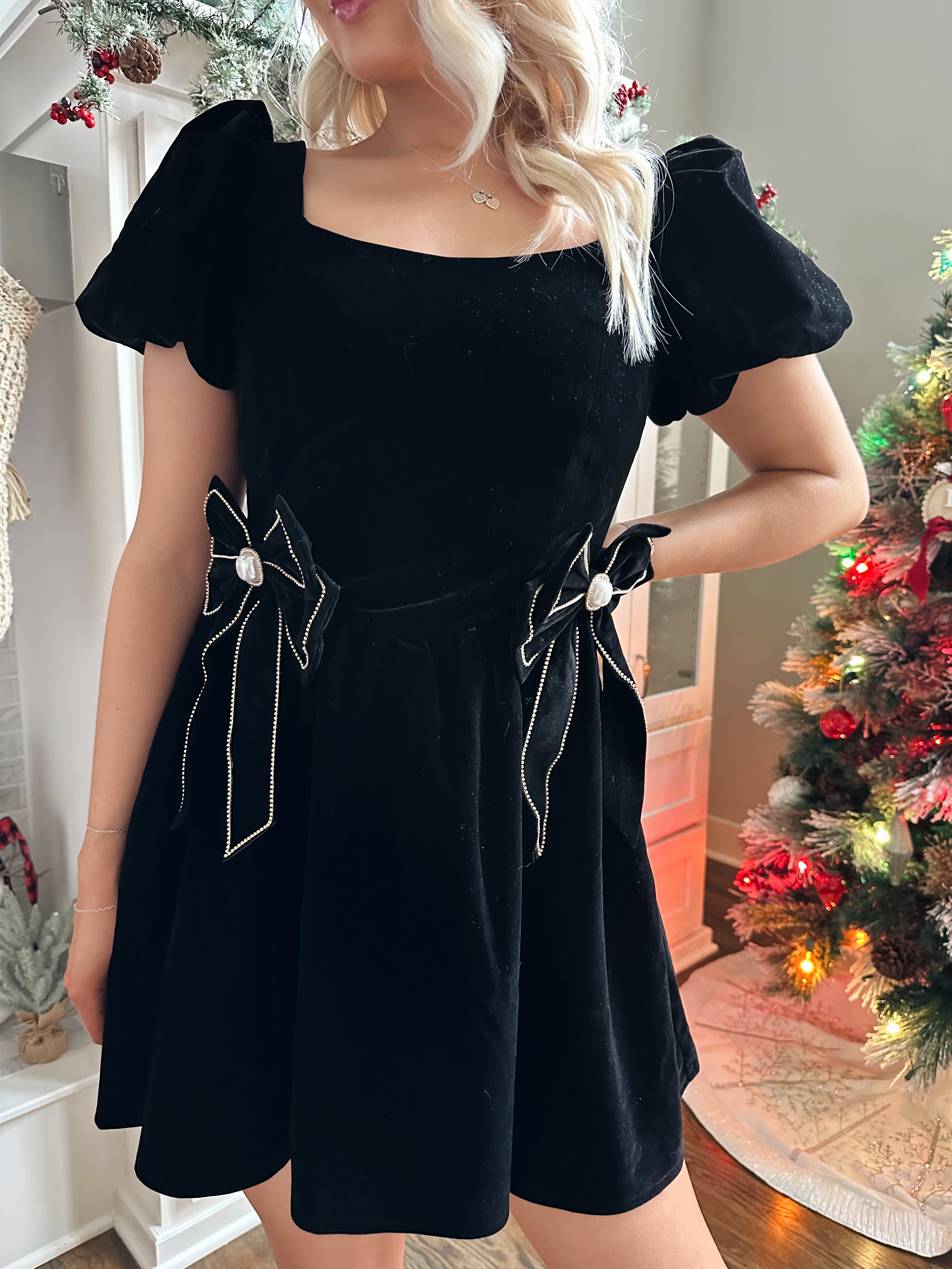 Woman wearing black velvet babydoll mini dress with puff sleeves, and bow detailing. 