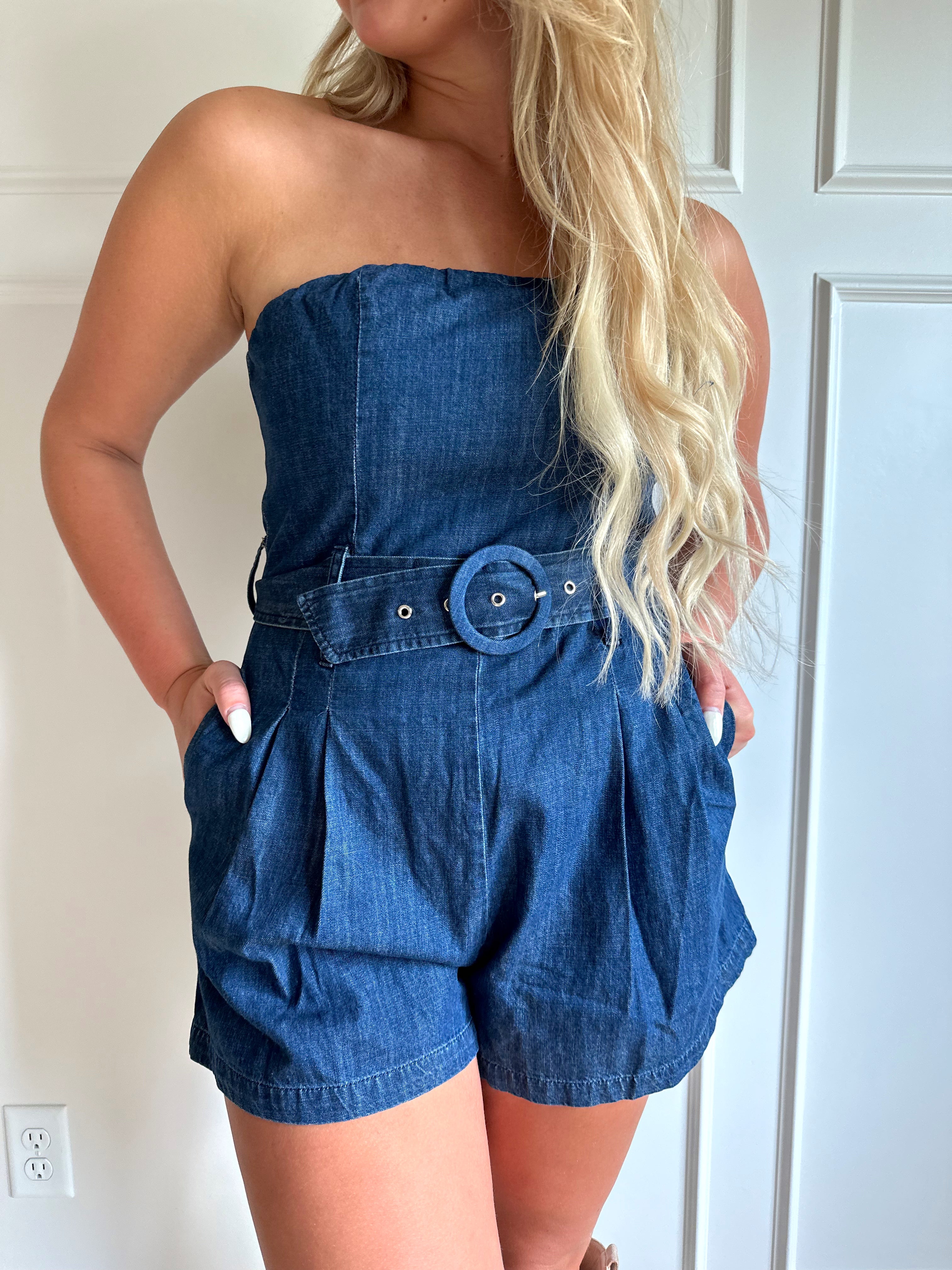 Woman wearing a dark wash strapless denim romper with a belted waist, and functional pockets.