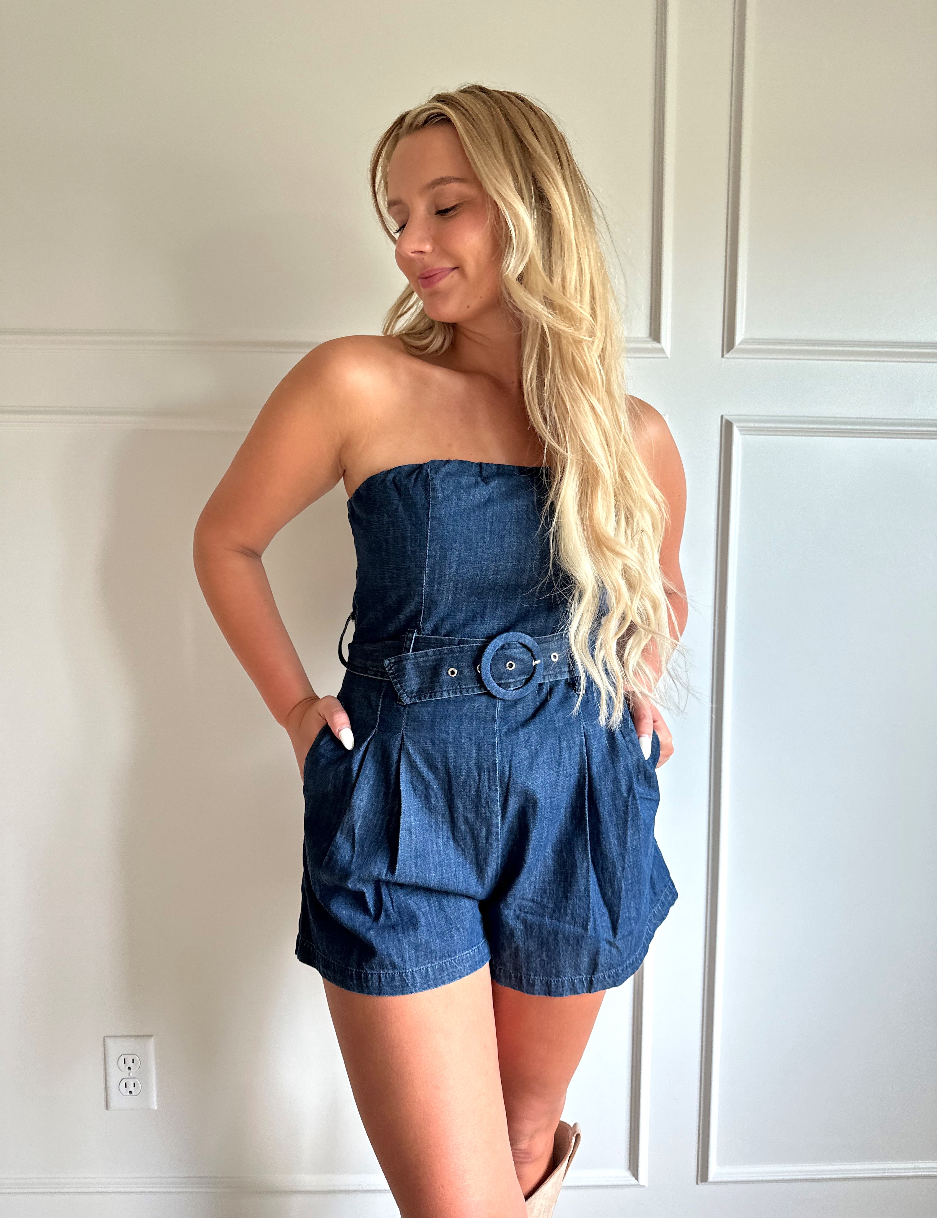 Woman wearing a dark wash strapless denim romper with a belted waist, and functional pockets.