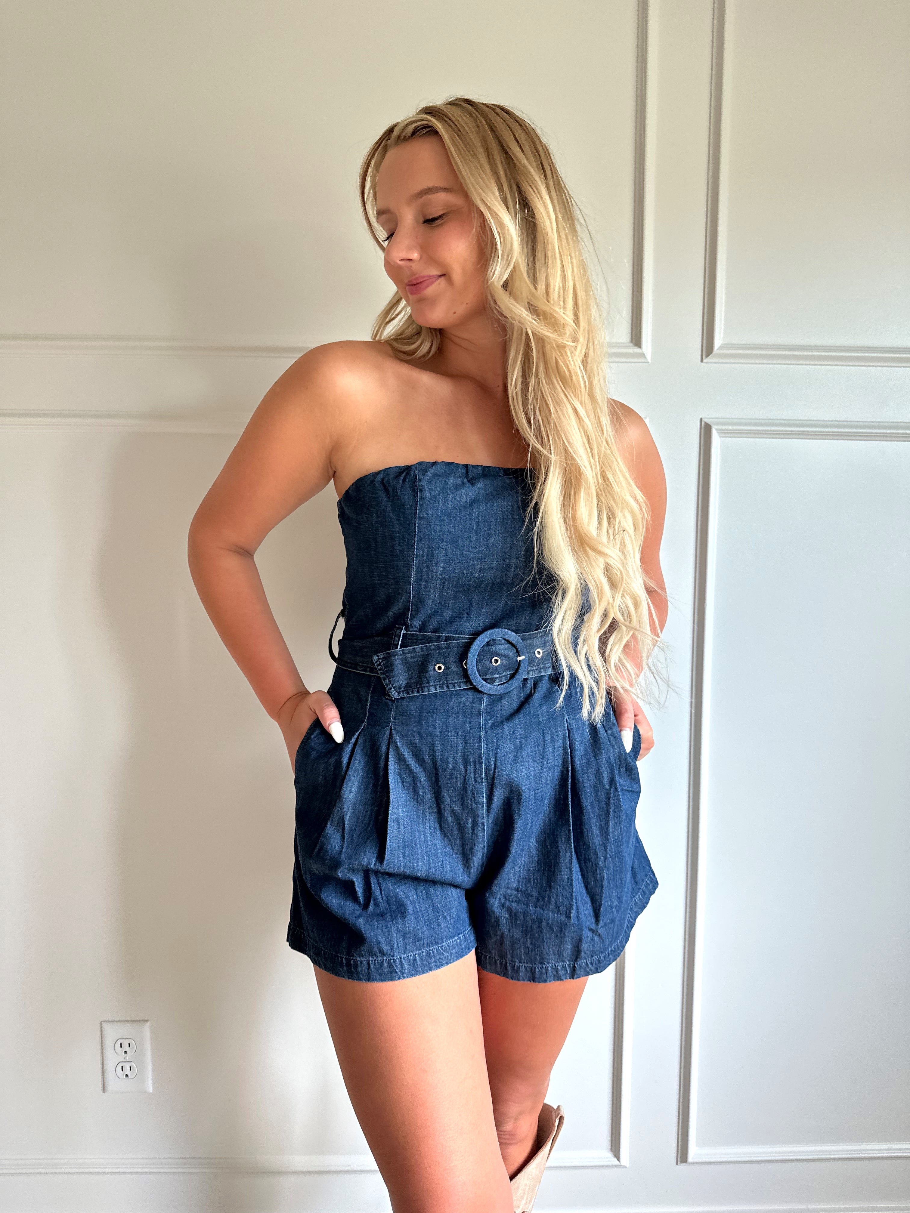 Woman wearing a dark wash strapless denim romper with a belted waist, and functional pockets.