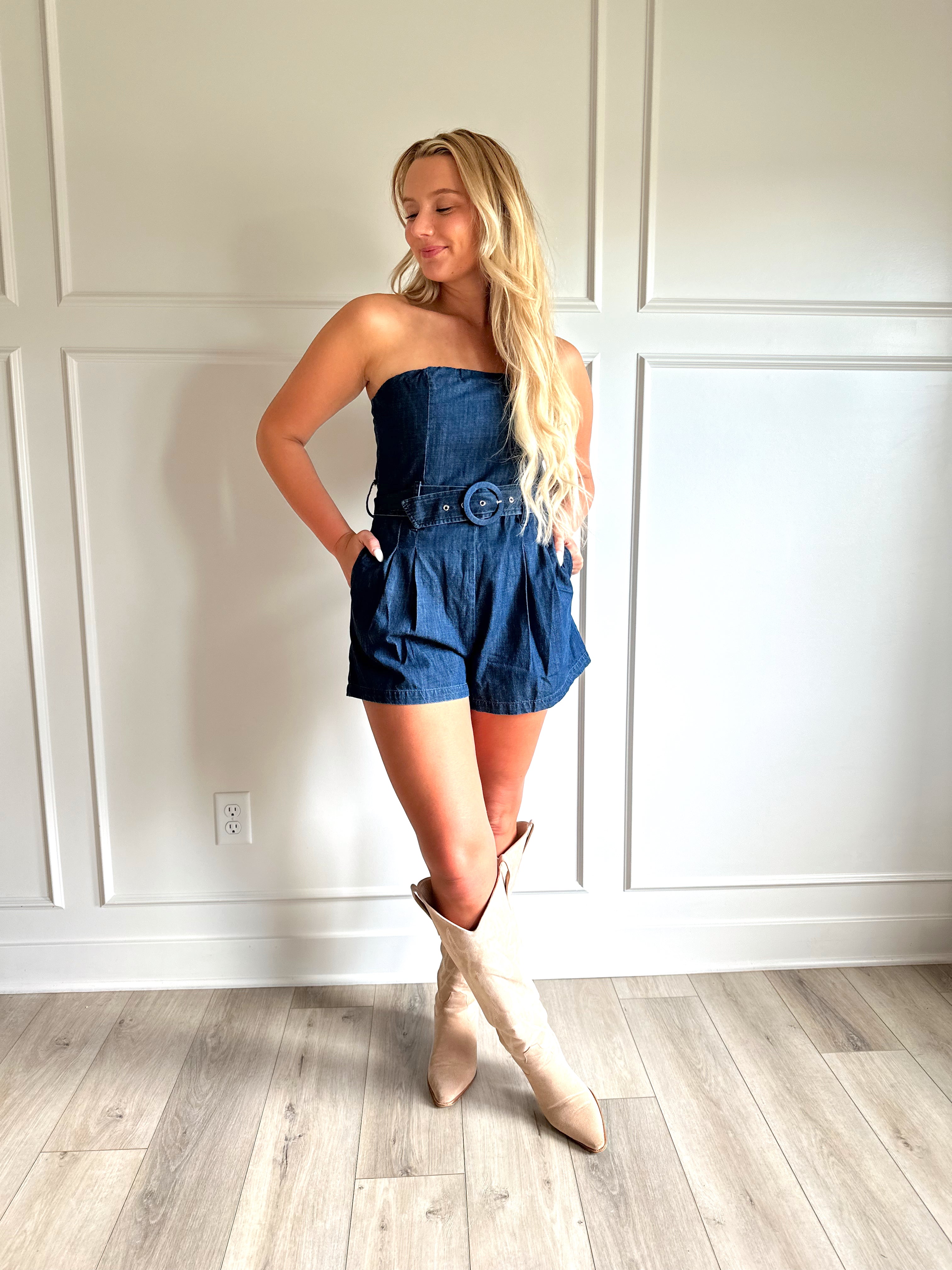 Woman wearing a dark wash strapless denim romper with a belted waist, and functional pockets.