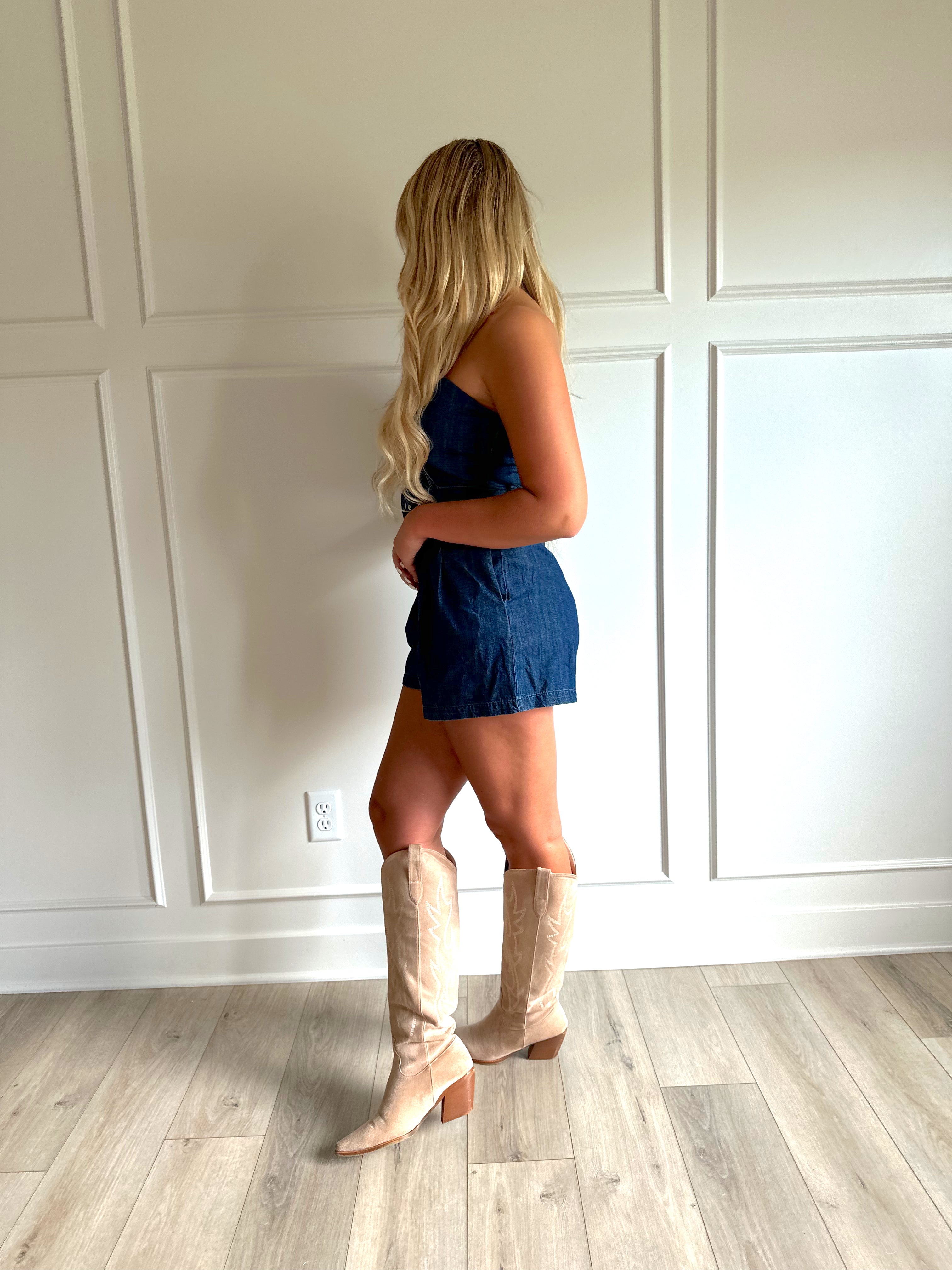 Woman wearing a dark wash strapless denim romper with a belted waist, and functional pockets.