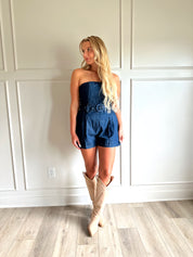 Woman wearing a dark wash strapless denim romper with a belted waist, and functional pockets.