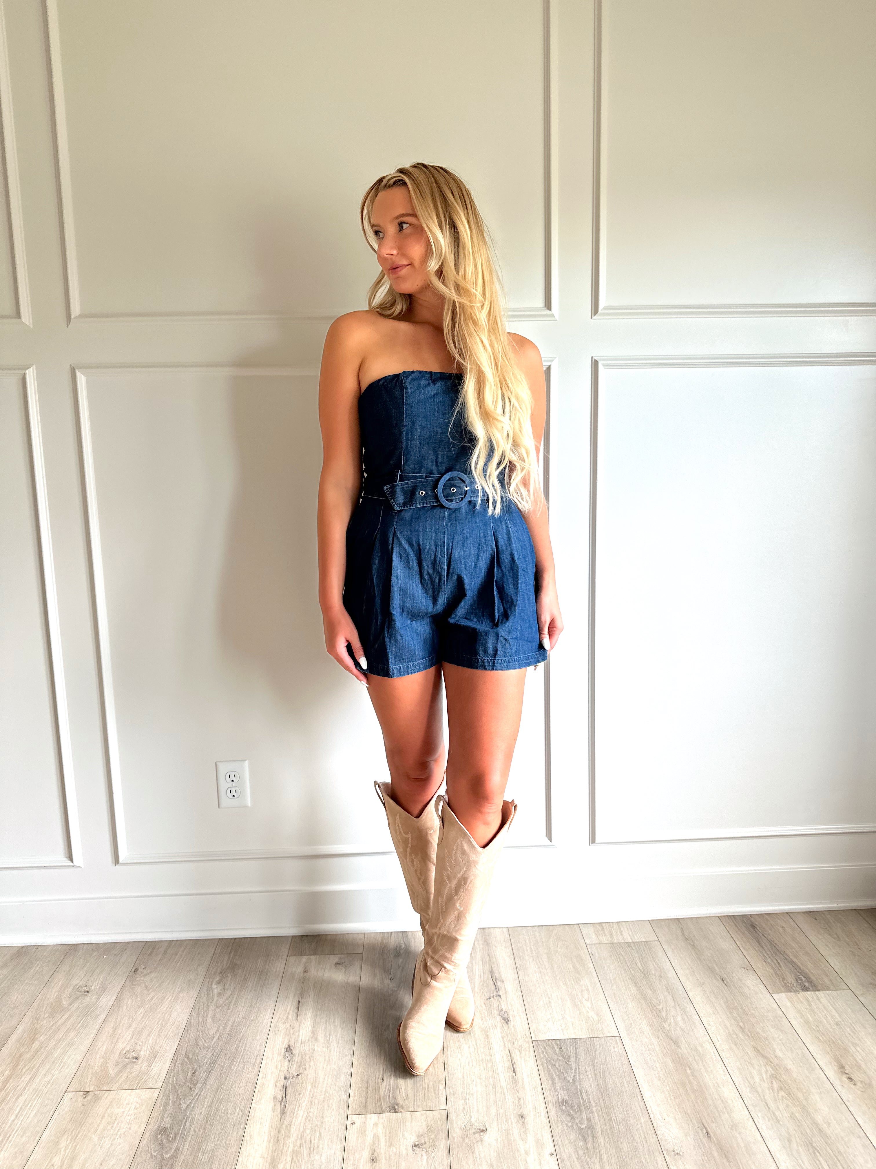Woman wearing a dark wash strapless denim romper with a belted waist, and functional pockets.
