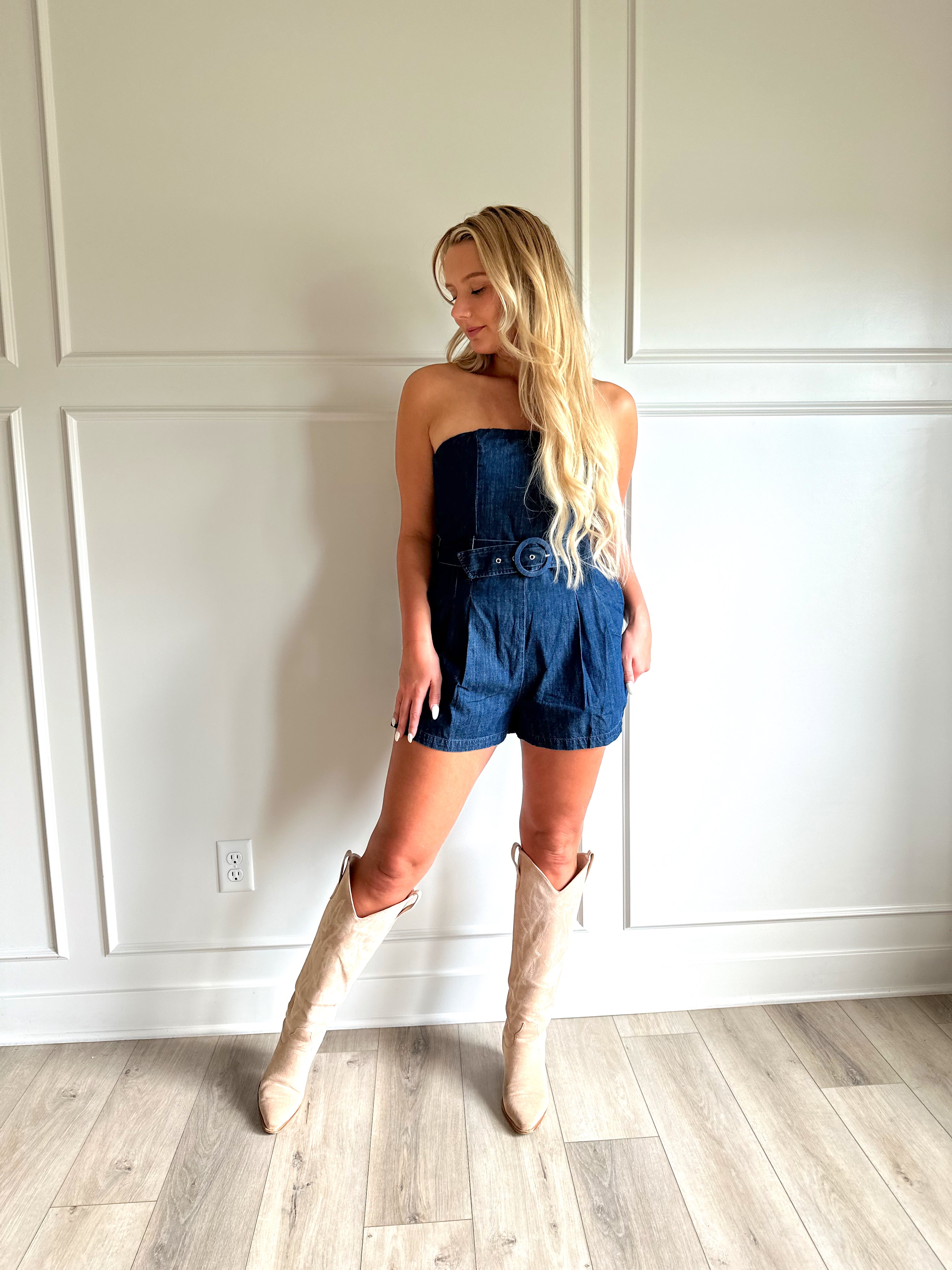 Woman wearing a dark wash strapless denim romper with a belted waist, and functional pockets.