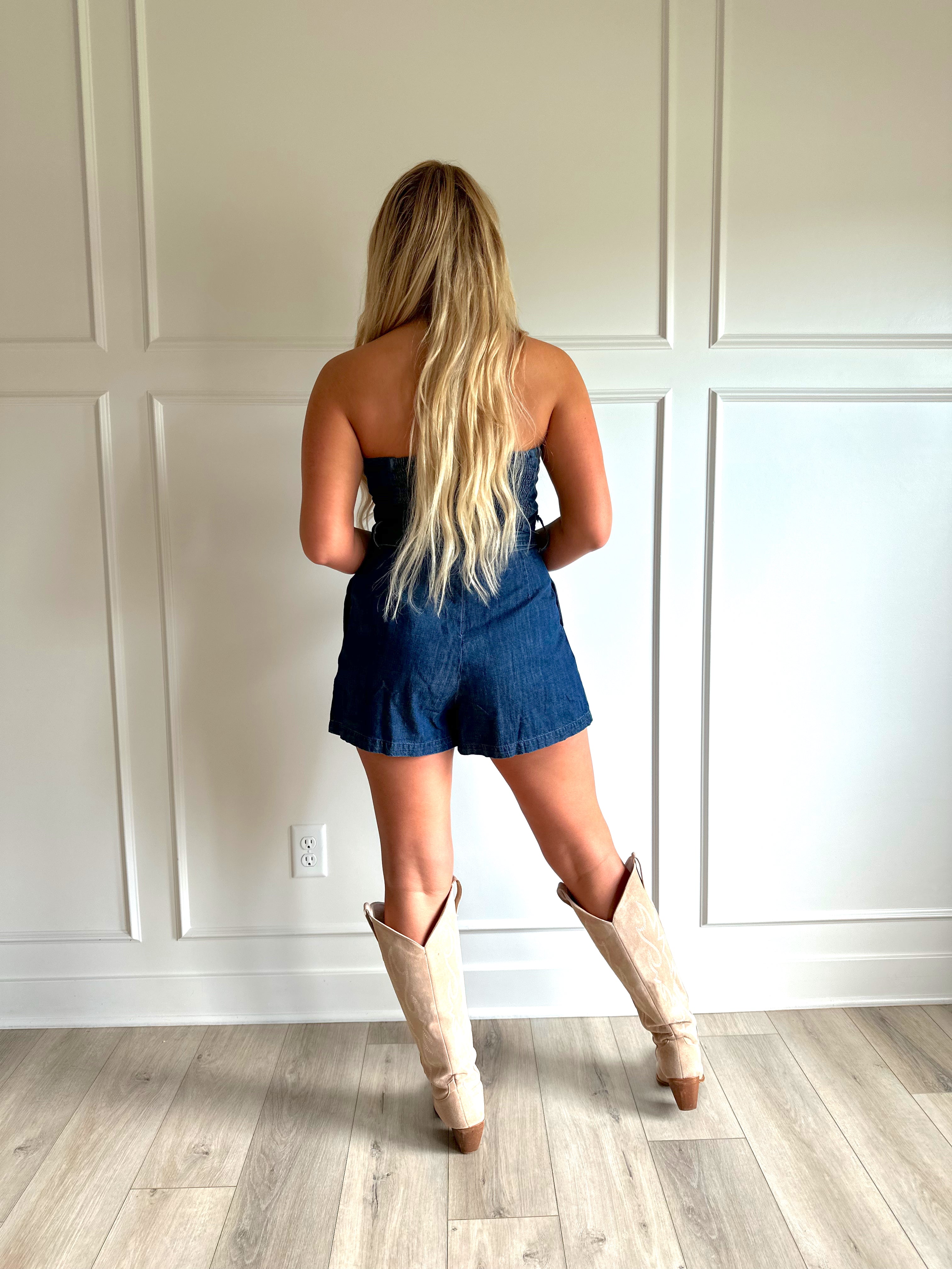 Woman wearing a dark wash strapless denim romper with a belted waist, and functional pockets.