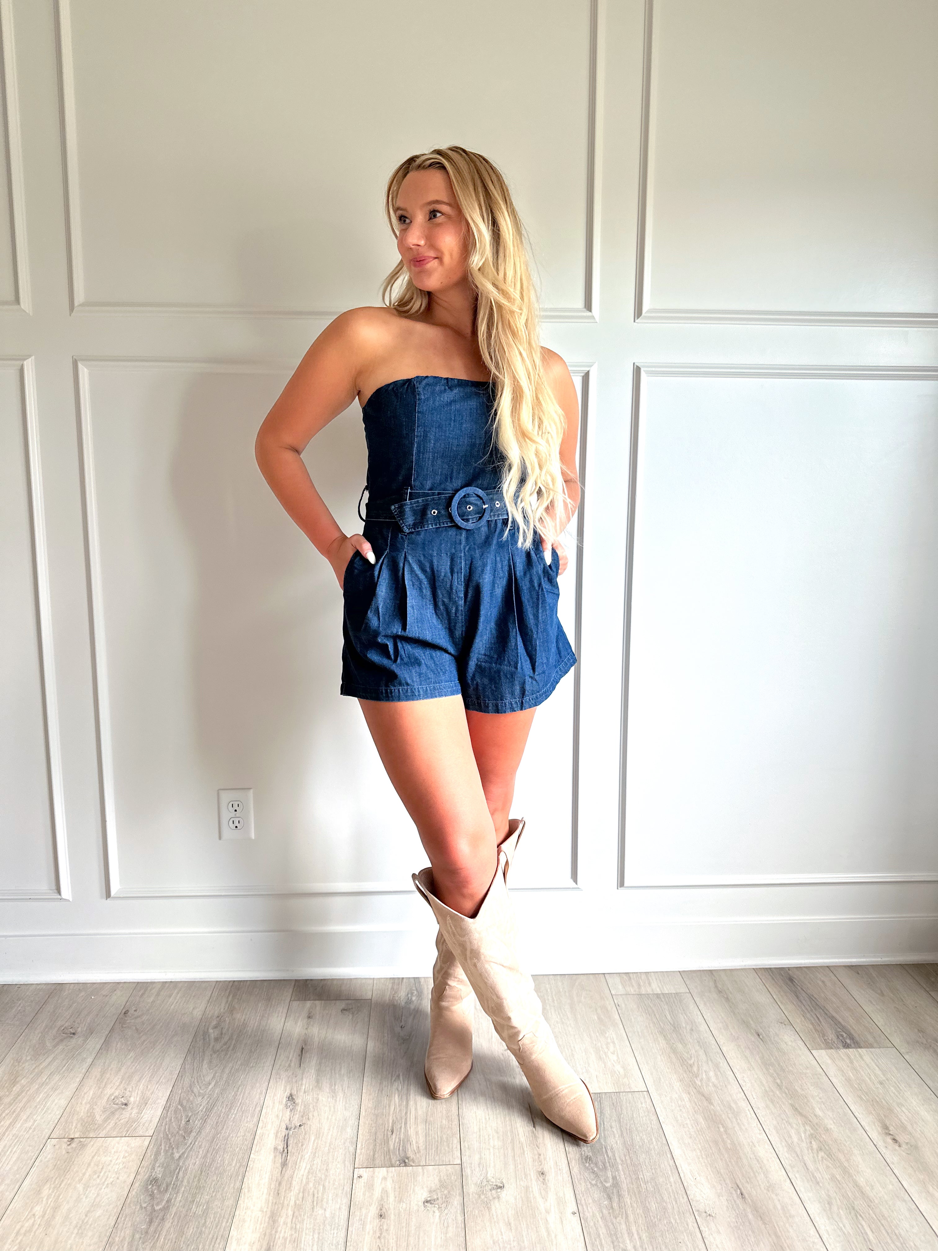 Woman wearing a dark wash strapless denim romper with a belted waist, and functional pockets.