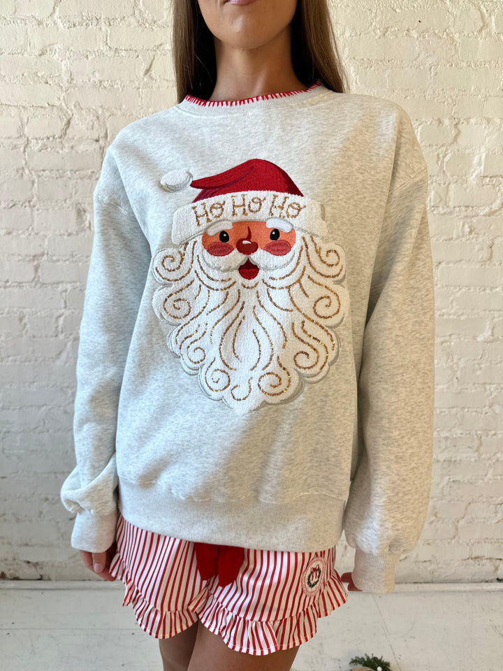 Woman wearing light grey pull over with an embroidered santa with sequin detail, and striped shorts. 