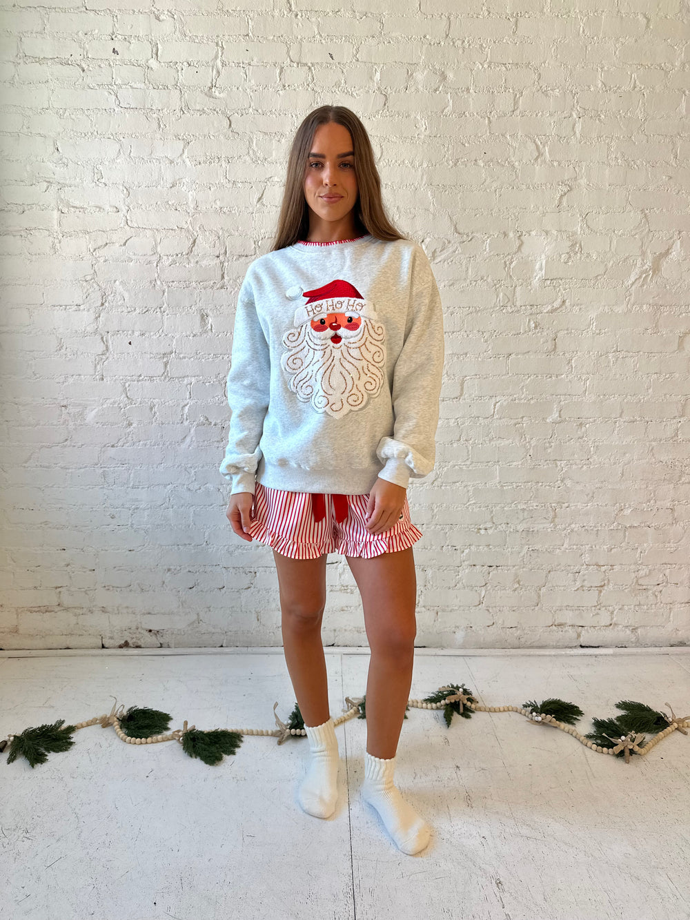 Woman wearing light grey pull over with an embroidered santa with sequin detail, and striped shorts. 