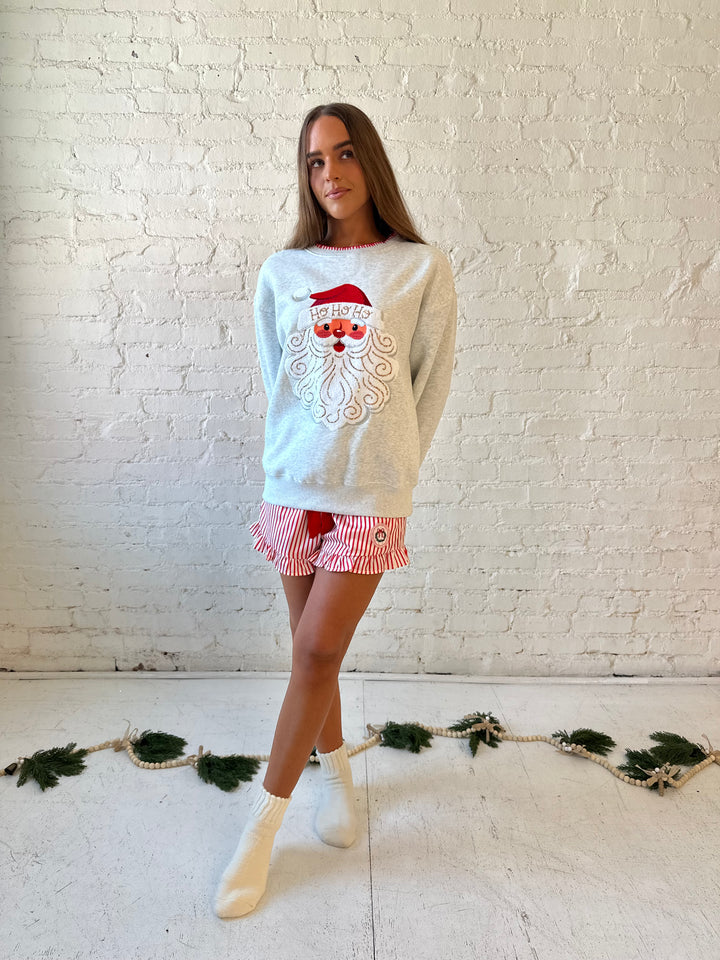 Woman wearing light grey pull over with an embroidered santa with sequin detail, and striped shorts. 