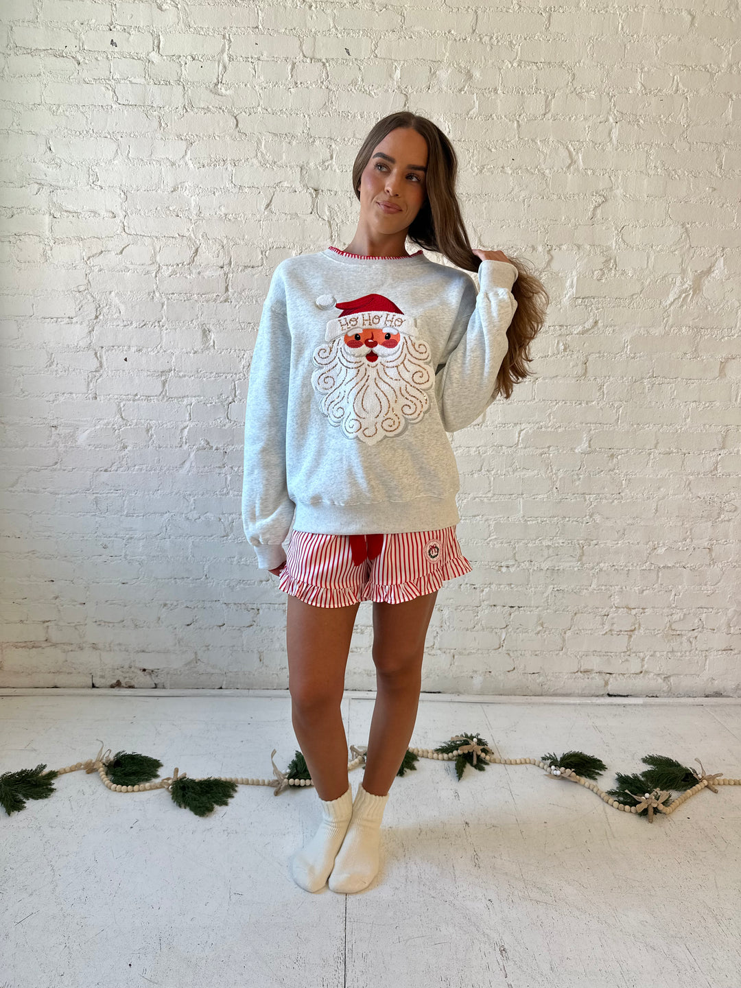 Woman wearing light grey pull over with an embroidered santa with sequin detail, and striped shorts. 