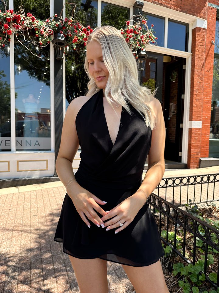 Woman wearing black wrap mini dress with self tie halter neck. 