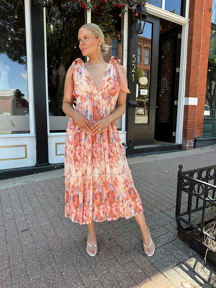 Woman wearing peach multi colored floral, pleated midi dress with v-neckline, and tie strap detail.