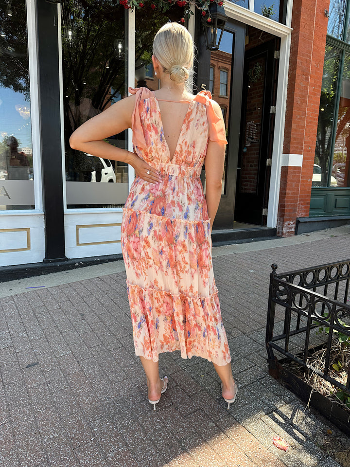 Woman wearing peach multi colored floral, pleated midi dress with v-neckline, and tie strap detail.