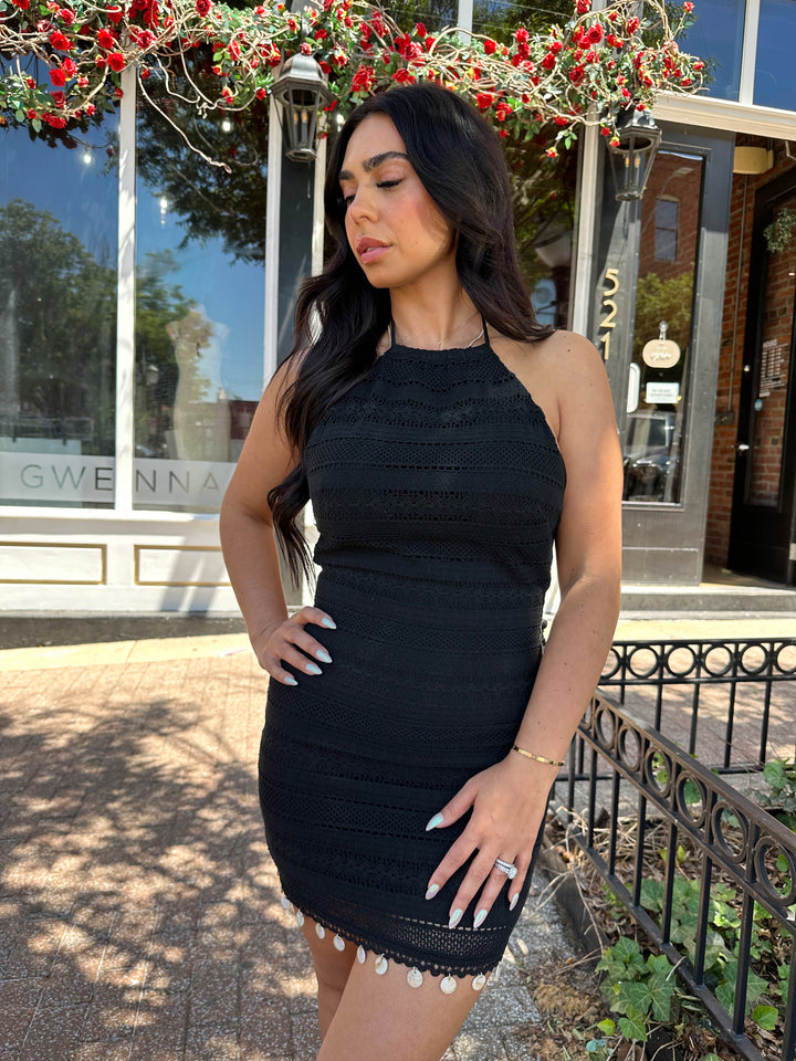 Woman wearing black crochet mini dress with beading hem detail, open back, and self tie straps. 
