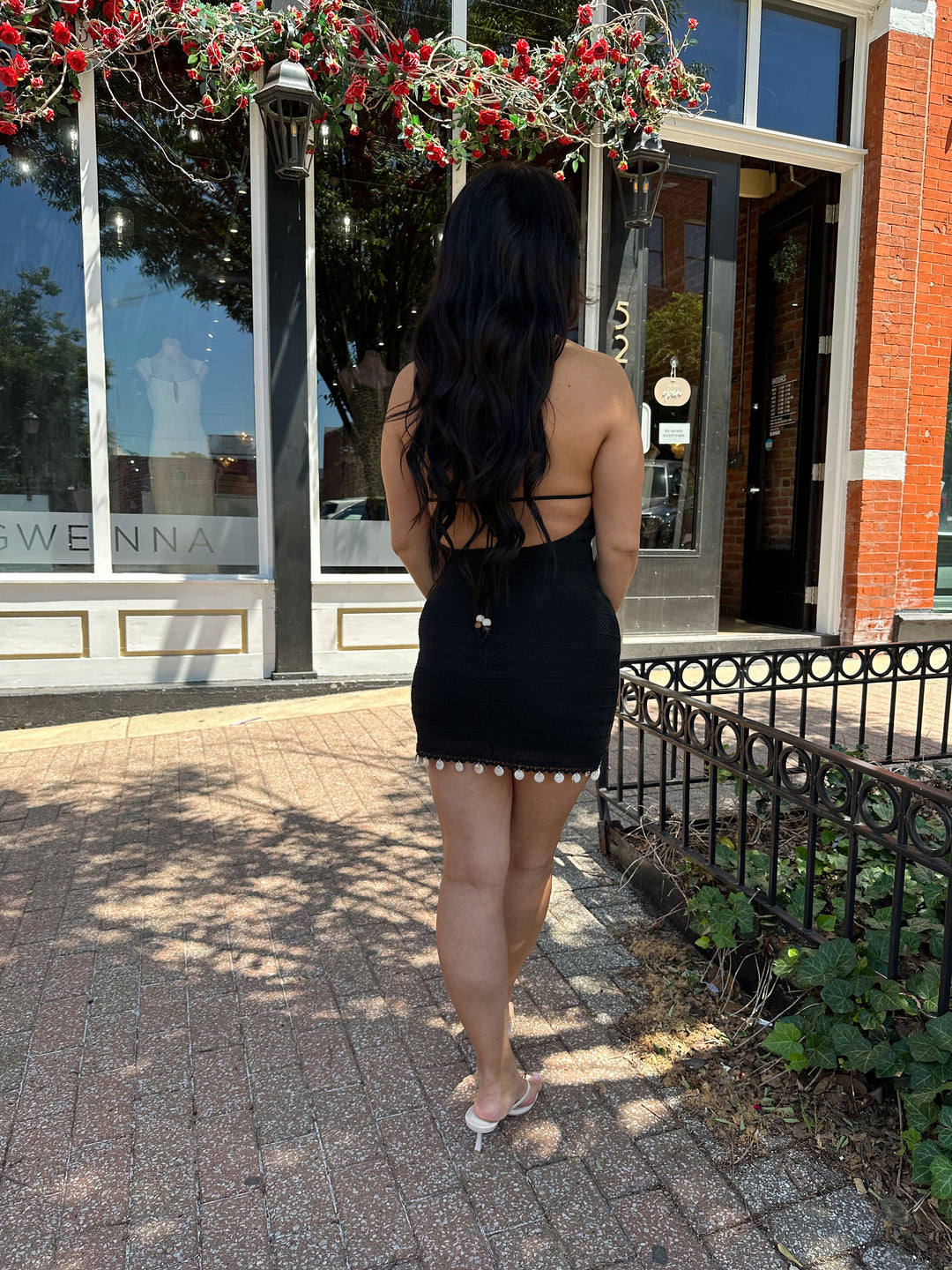 Woman wearing black crochet mini dress with beading hem detail, open back, and self tie straps. 