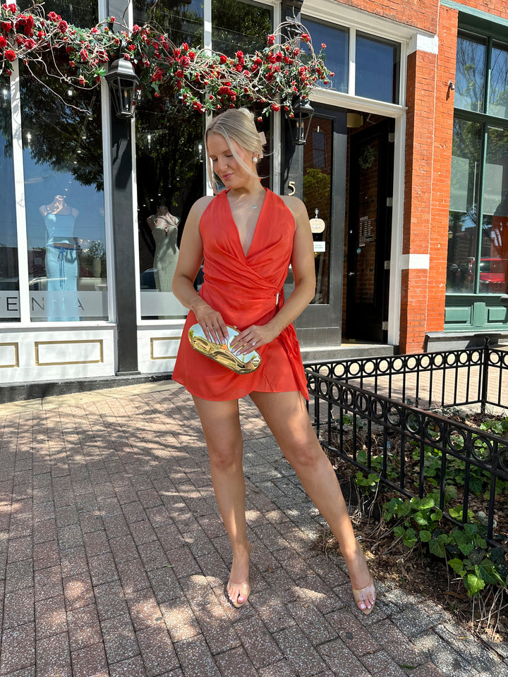 Woman wearing light weight color wrap mini dress with self tie halter neck, and gold hardware.
