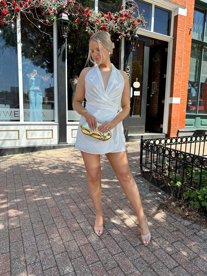 Woman wearing light weight white wrap mini dress with self tie halter neck, and gold hardware. 