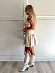 Woman wearing light beige drop waist mini dress with bow detail and cream knee boot.