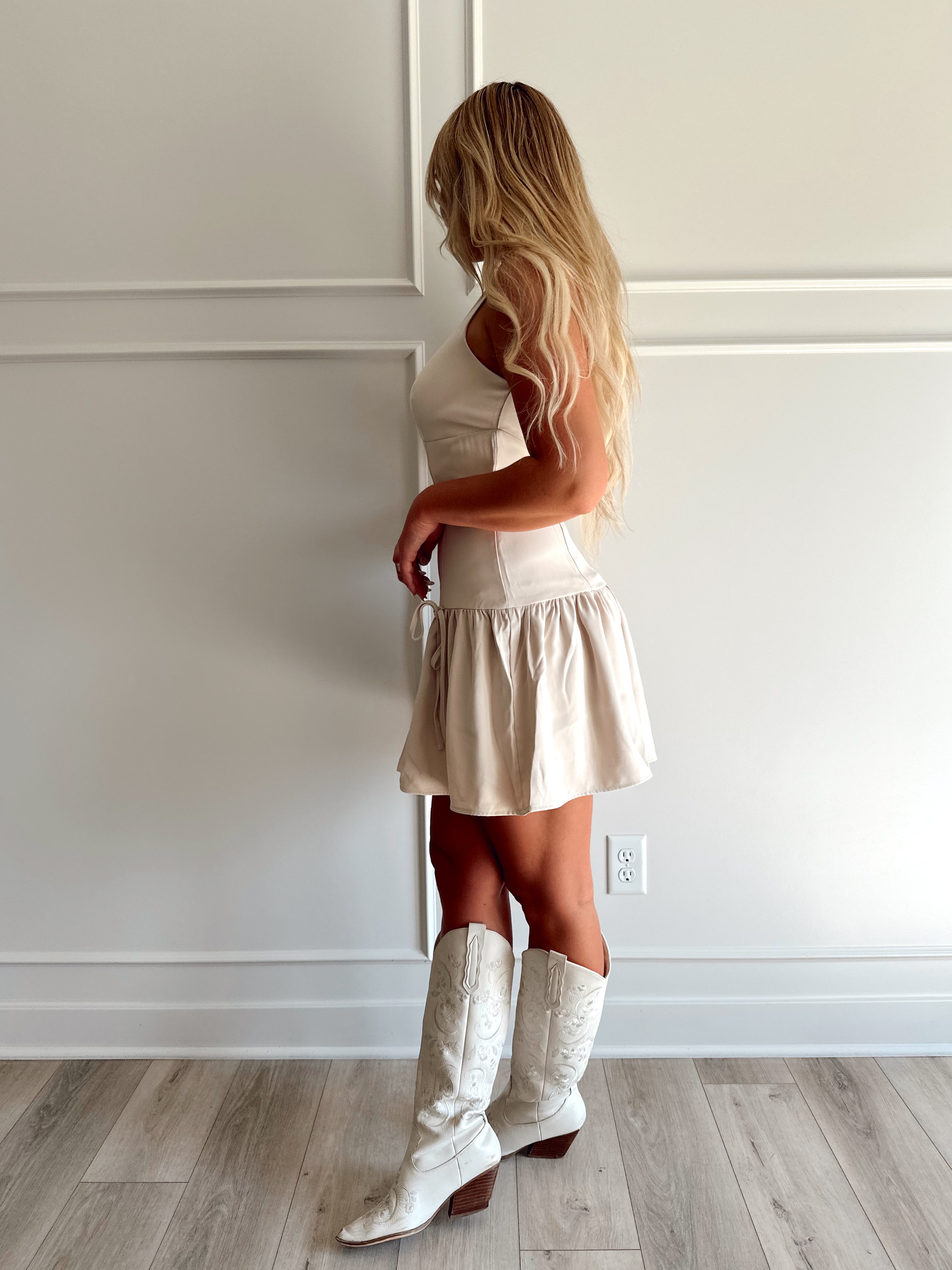 Woman wearing light beige drop waist mini dress with bow detail and cream knee boot.
