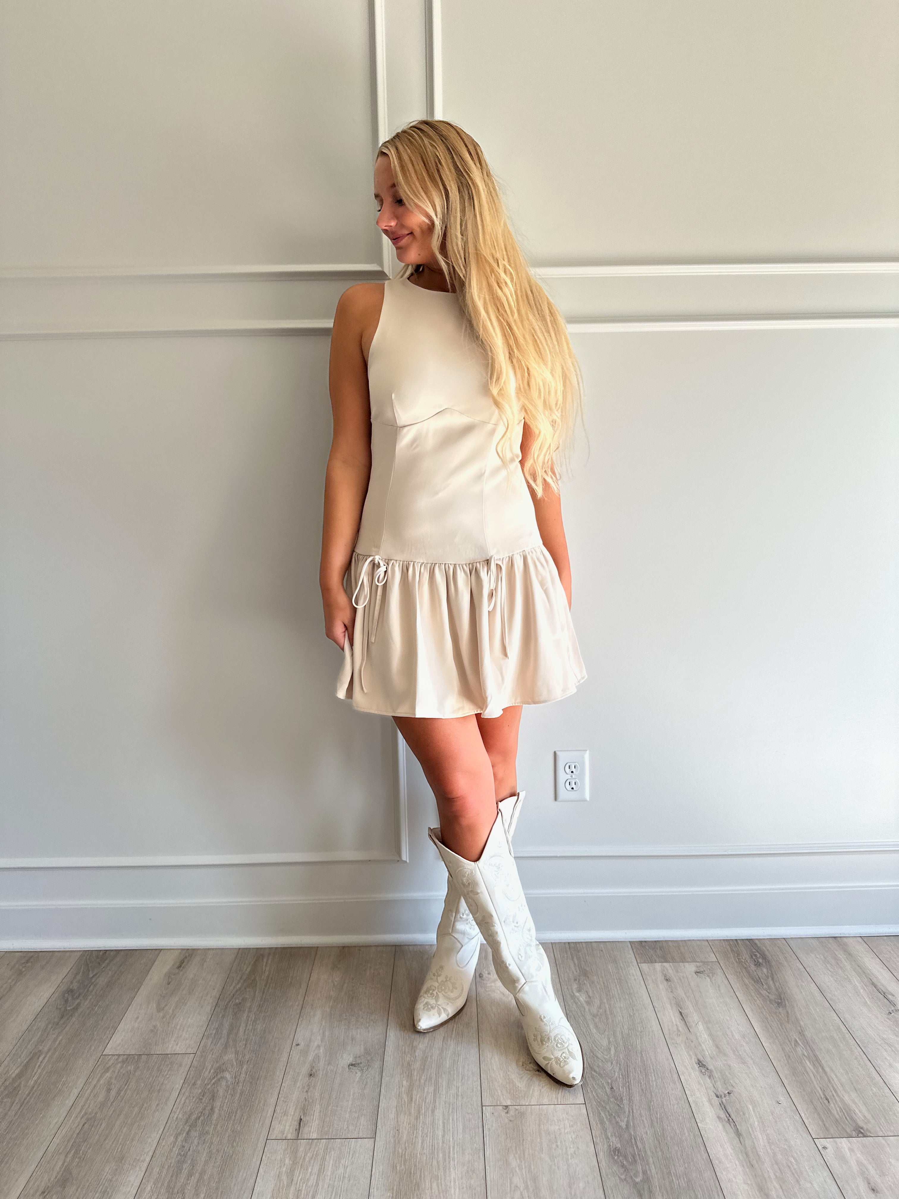 Woman wearing light beige drop waist mini dress with bow detail and cream knee boot.