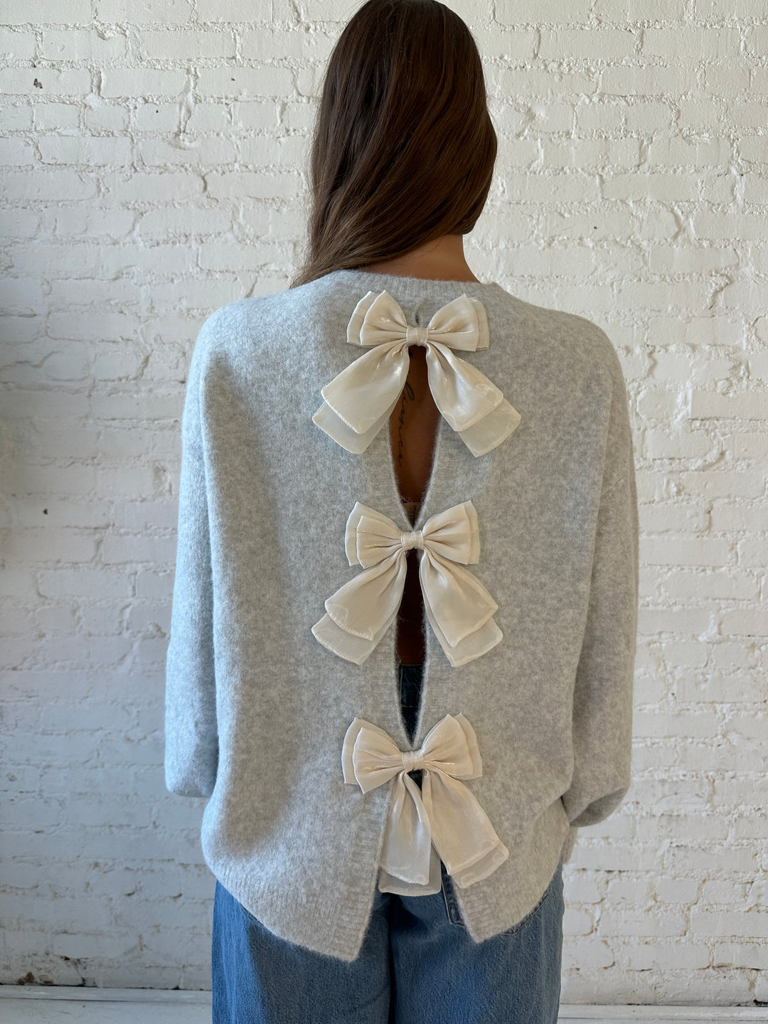 Woman wearing grey knit sweater with cream bow back detail with medium wash jeans. 