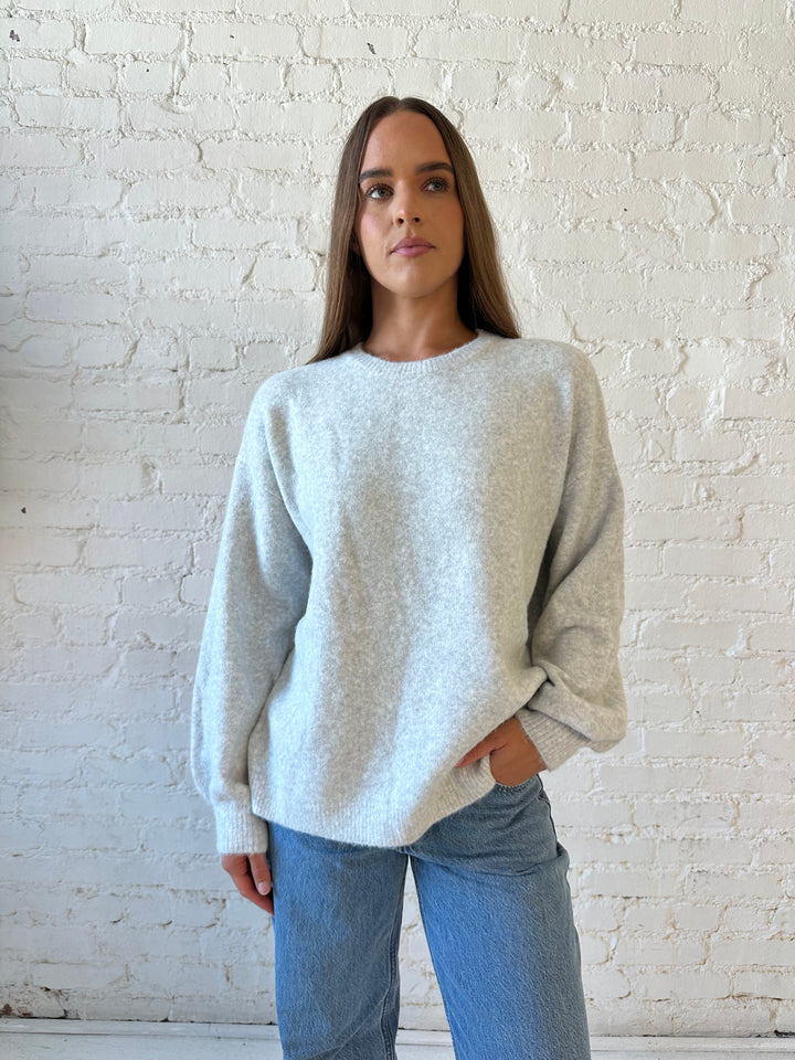 Woman wearing grey knit sweater with cream bow back detail with medium wash jeans. 