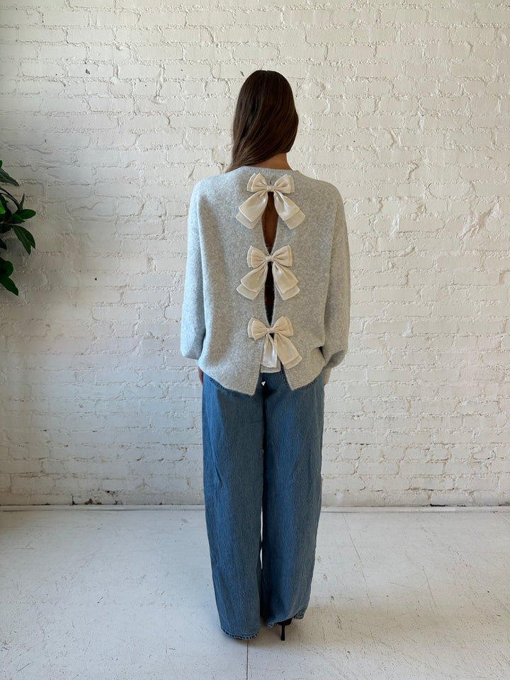 Woman wearing grey knit sweater with cream bow back detail with medium wash jeans. 