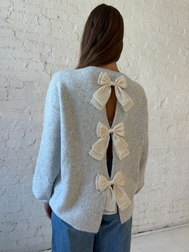 Woman wearing grey knit sweater with cream bow back detail with medium wash jeans. 