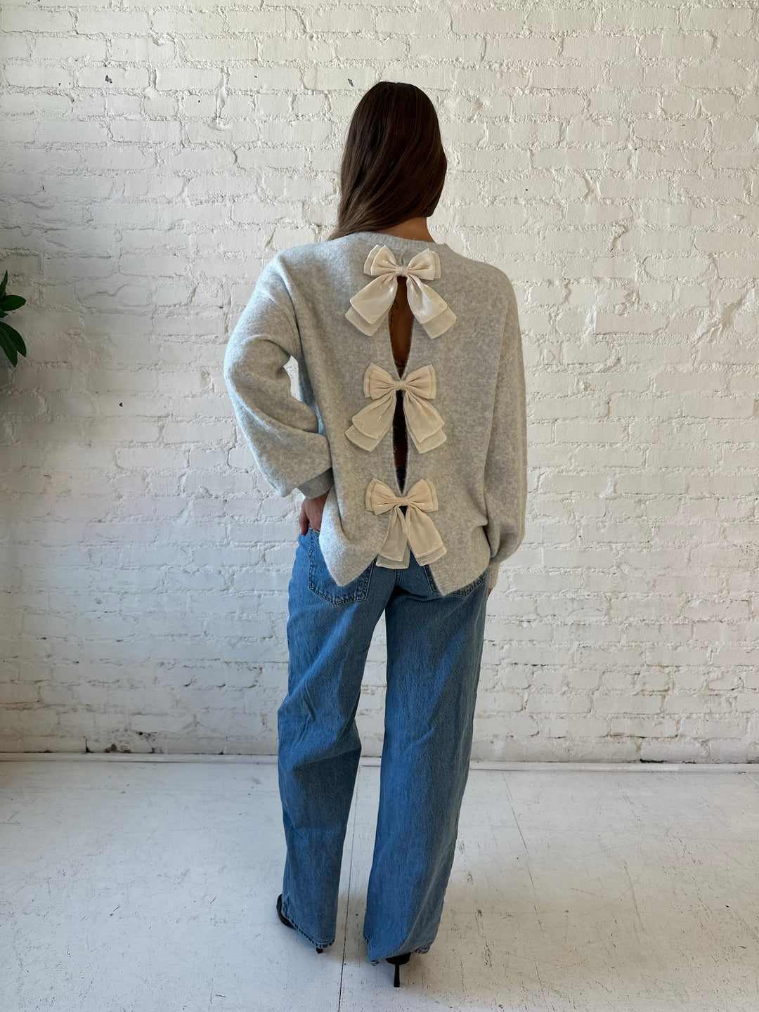 Woman wearing grey knit sweater with cream bow back detail with medium wash jeans. 