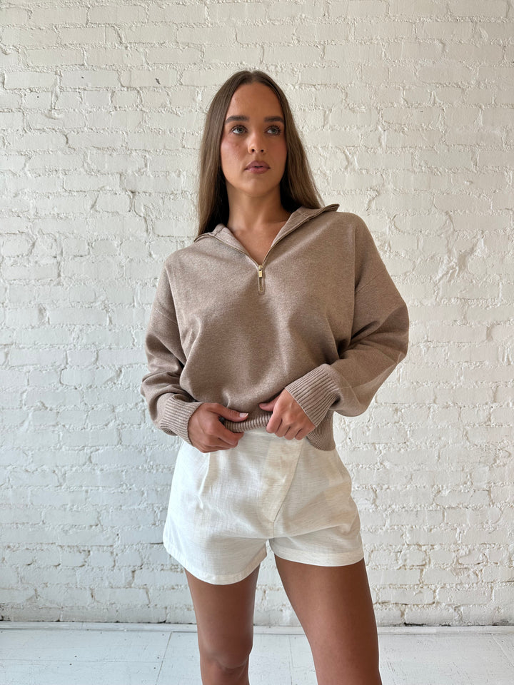 Woman wearing tan knit quarter zip sweater with cream shorts.
