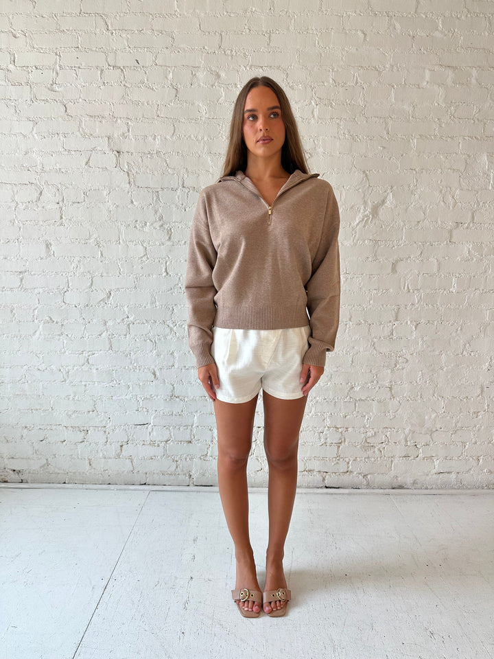 Woman wearing tan knit quarter zip sweater with cream shorts.
