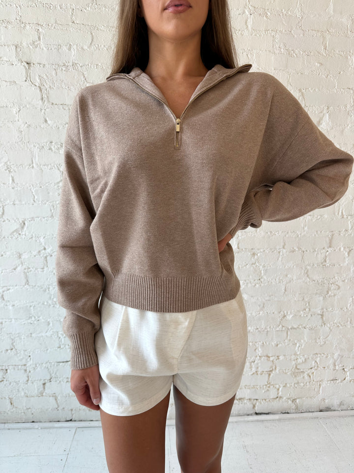 Woman wearing tan knit quarter zip sweater with cream shorts.