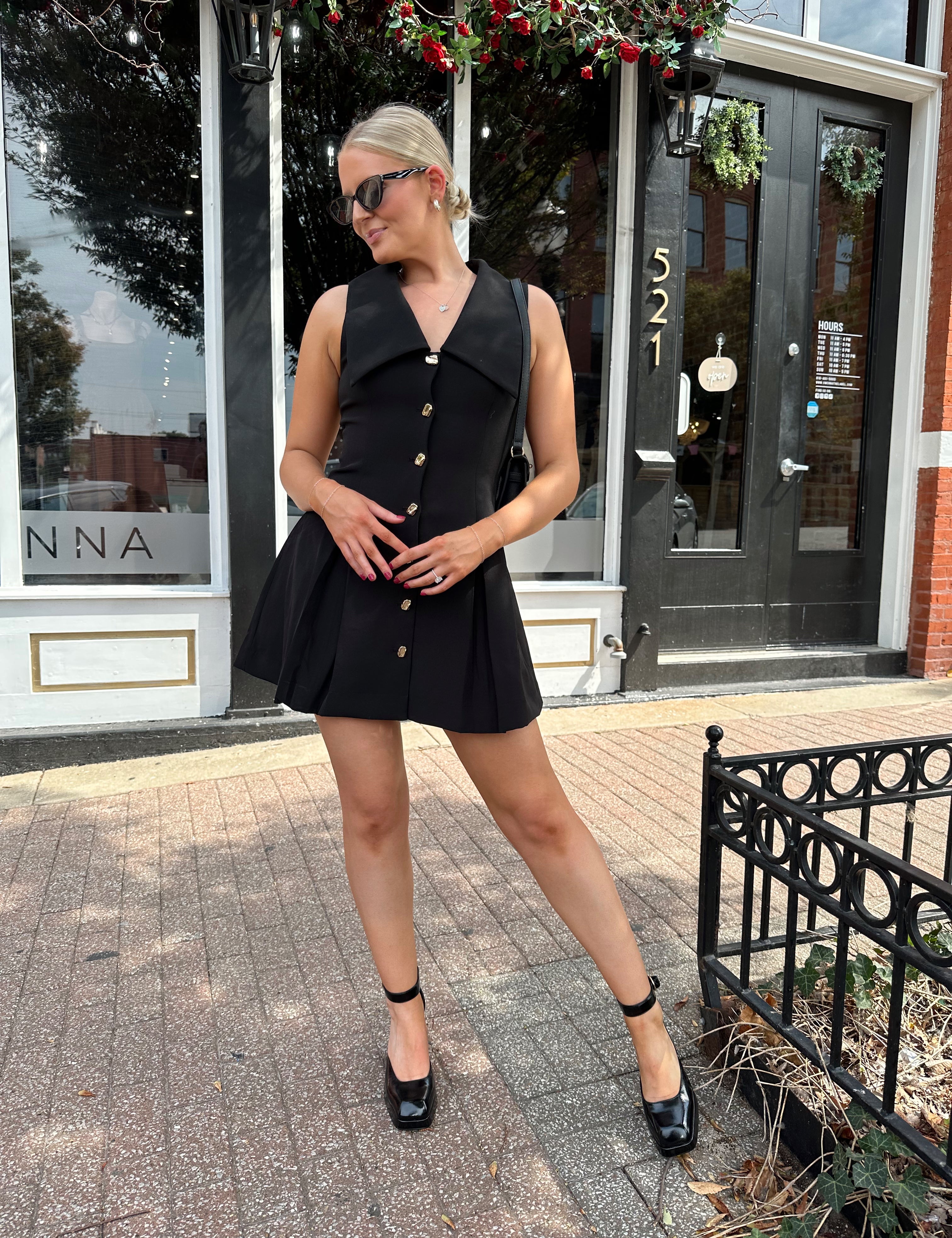 Woman wearing black tailored mini dress with oversized collar neckline and gold button closure. 