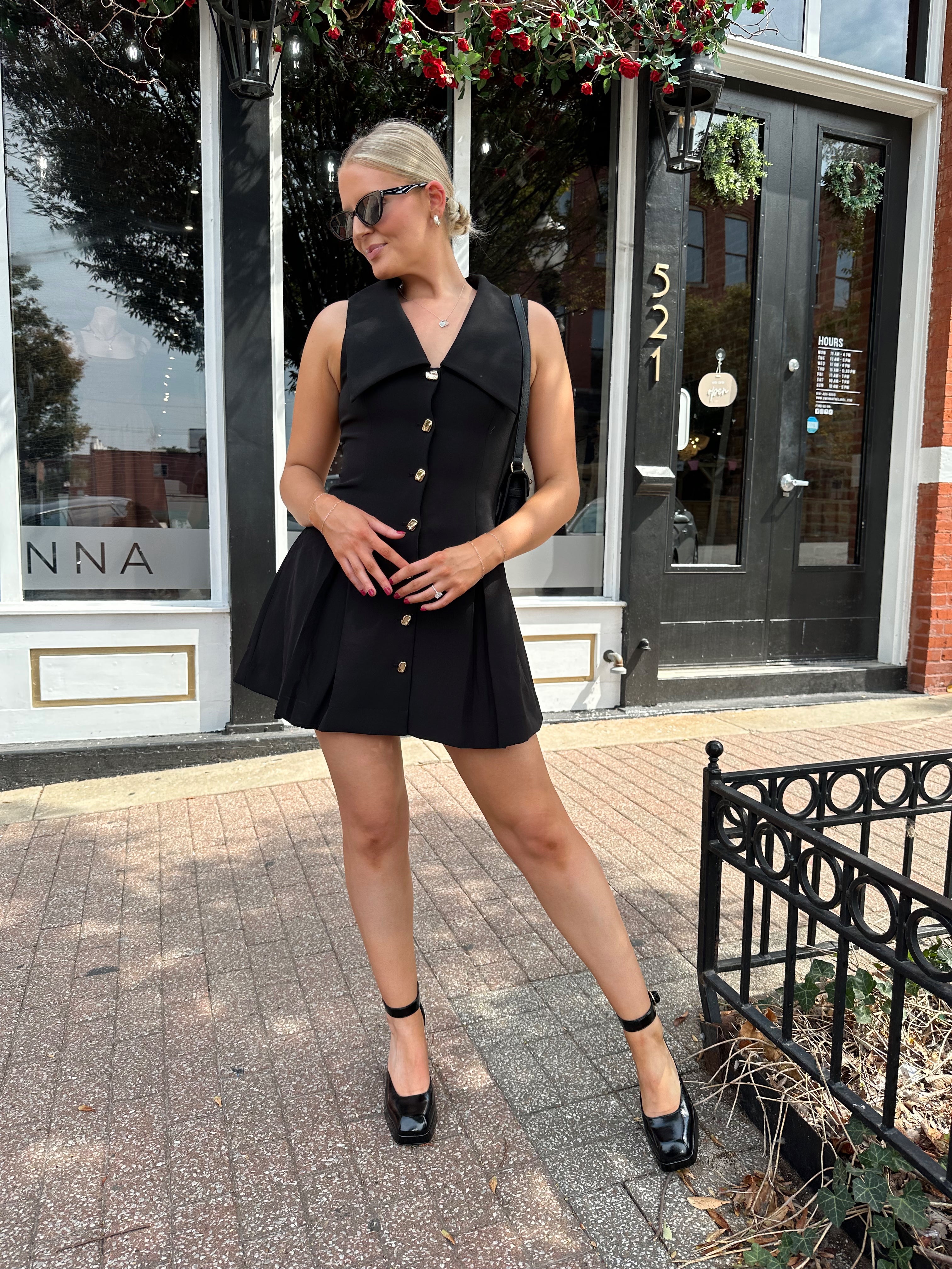 Woman wearing black tailored mini dress with oversized collar neckline and gold button closure. 