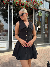 Woman wearing black tailored mini dress with oversized collar neckline and gold button closure. 
