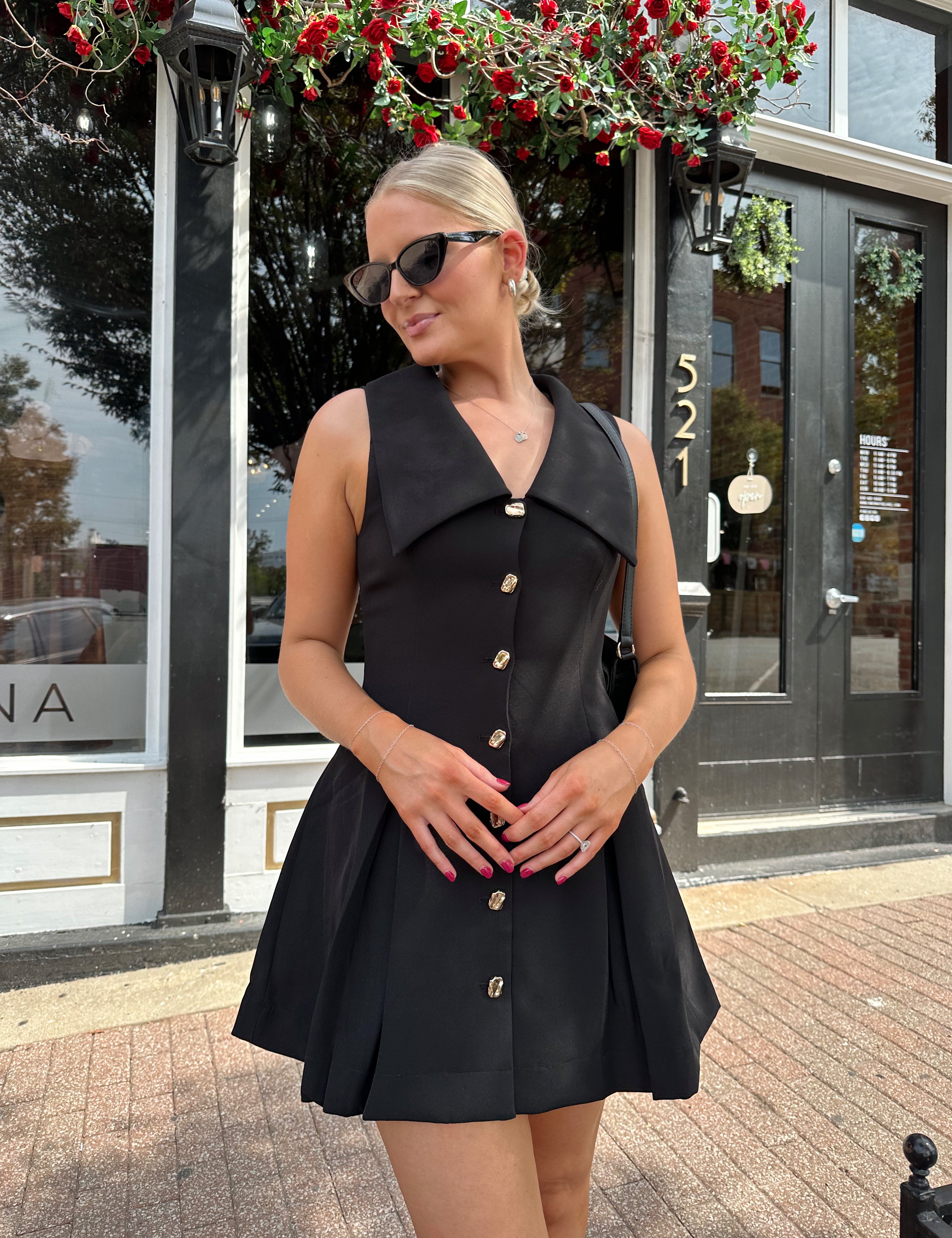 Woman wearing black tailored mini dress with oversized collar neckline and gold button closure. 