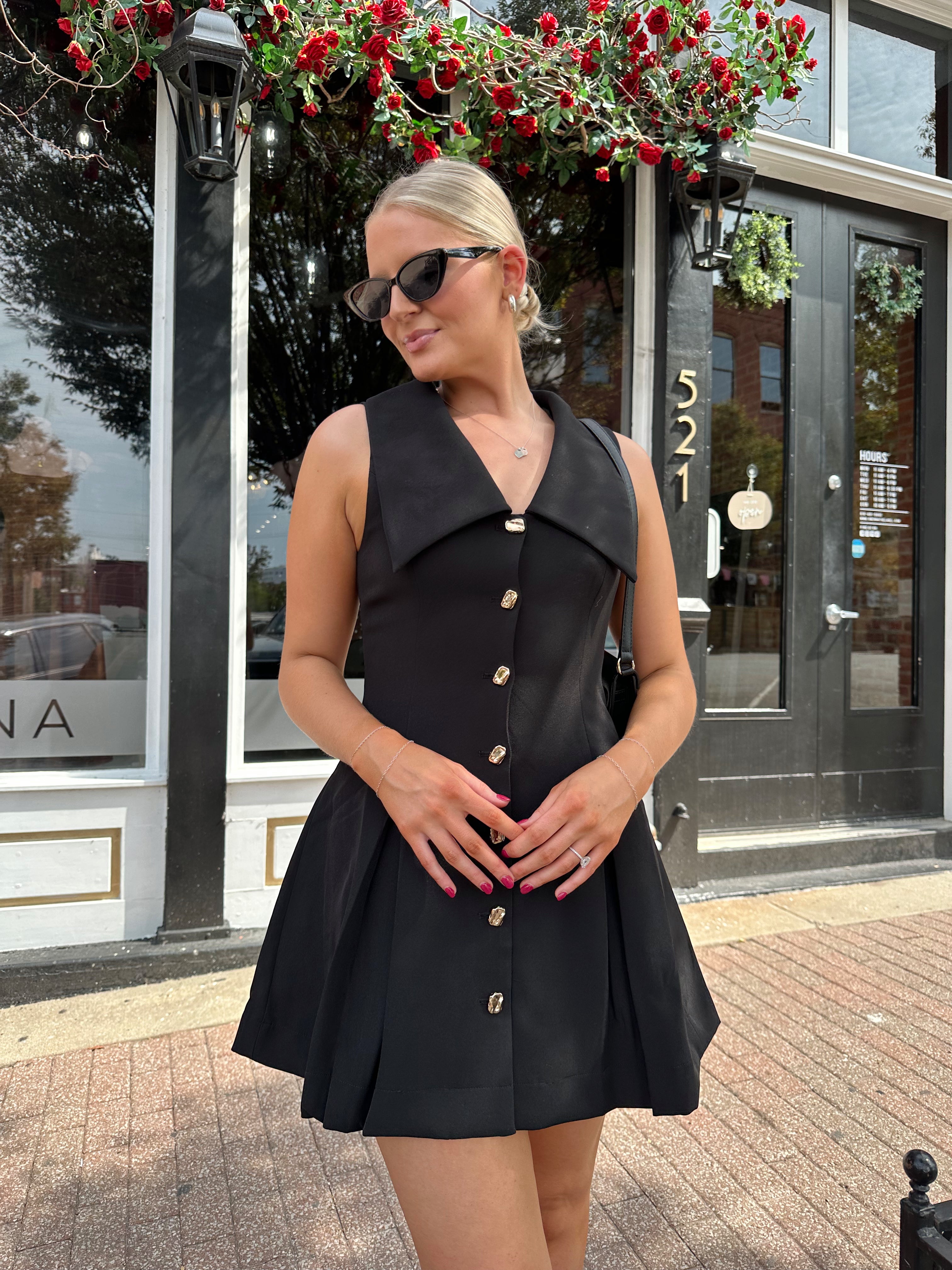 Woman wearing black tailored mini dress with oversized collar neckline and gold button closure. 