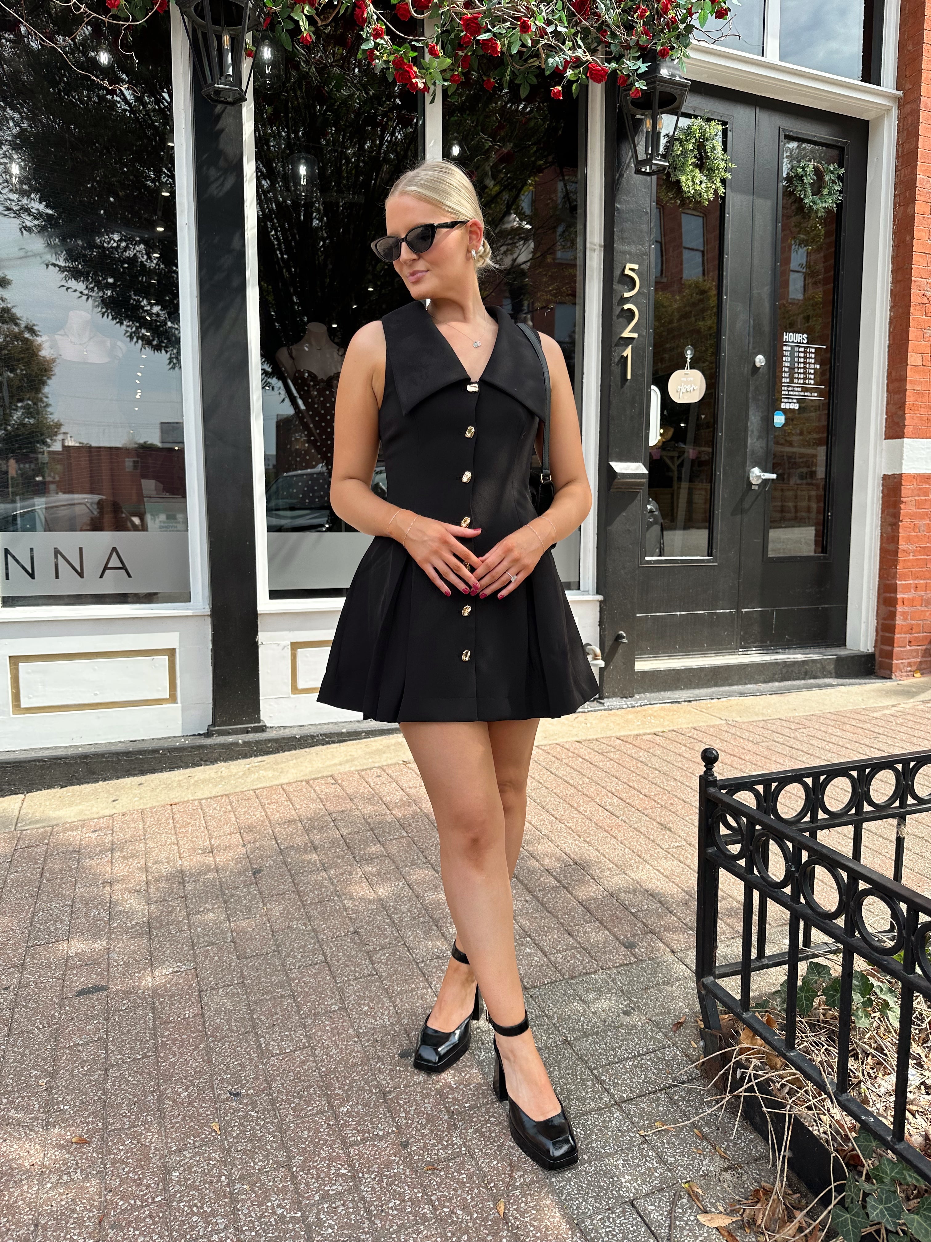 Woman wearing black tailored mini dress with oversized collar neckline and gold button closure. 