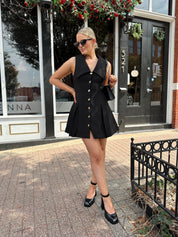 Woman wearing black tailored mini dress with oversized collar neckline and gold button closure. 
