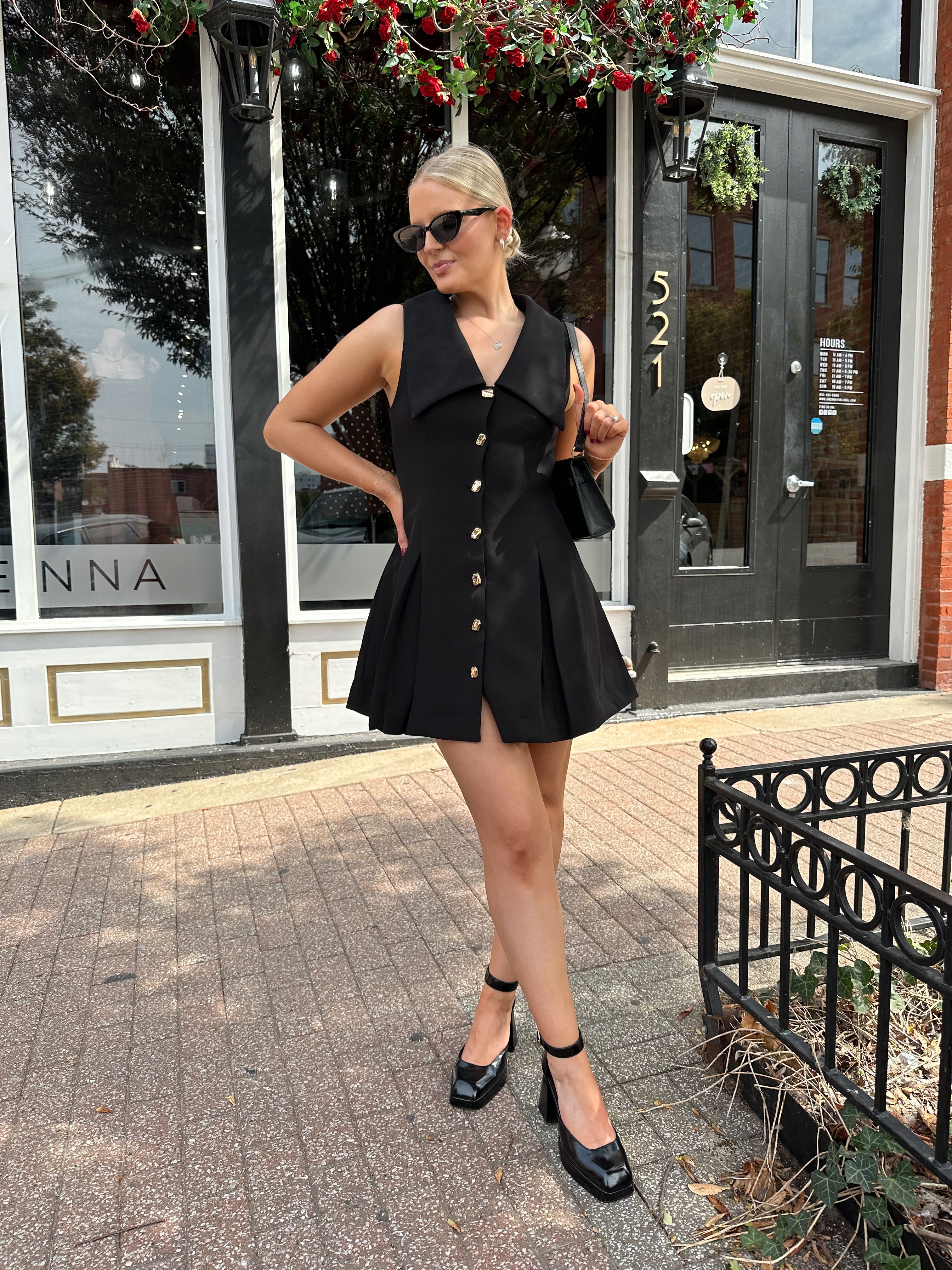 Woman wearing black tailored mini dress with oversized collar neckline and gold button closure. 