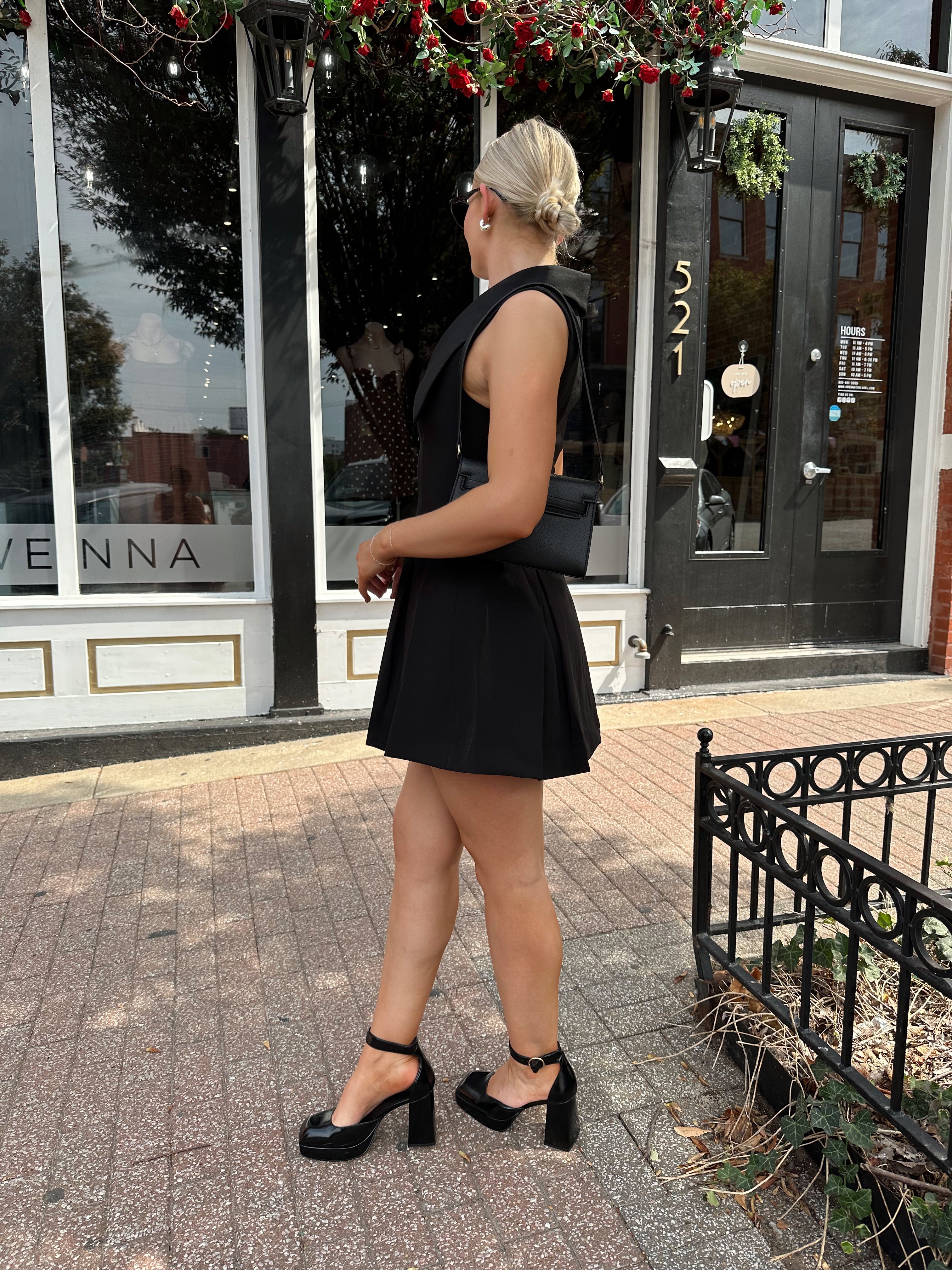 Woman wearing black tailored mini dress with oversized collar neckline and gold button closure. 