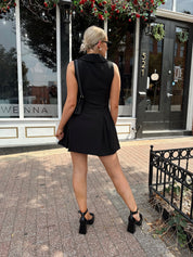 Woman wearing black tailored mini dress with oversized collar neckline and gold button closure. 