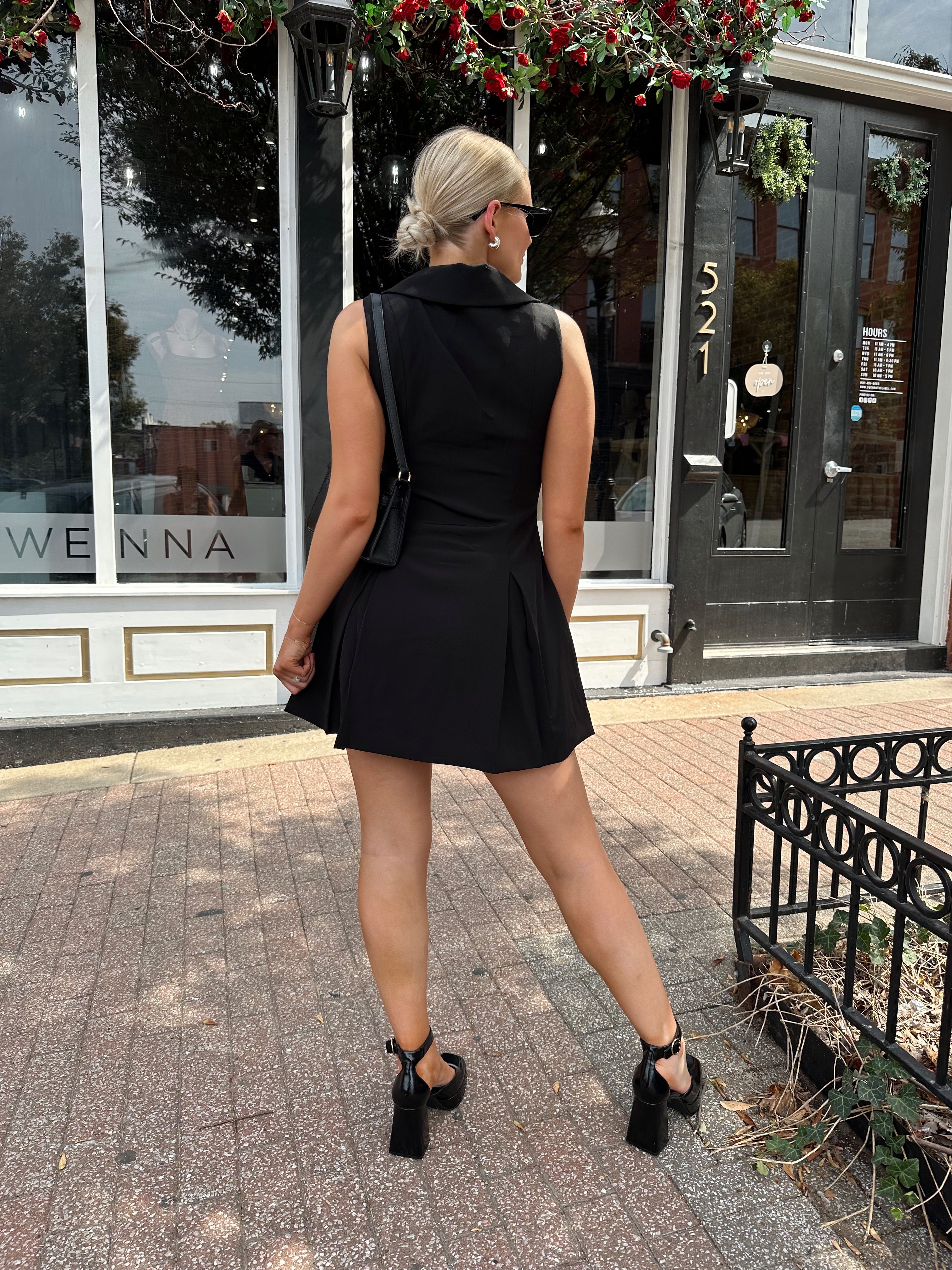 Woman wearing black tailored mini dress with oversized collar neckline and gold button closure. 