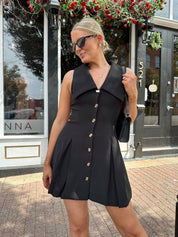 Woman wearing black tailored mini dress with oversized collar neckline and gold button closure. 