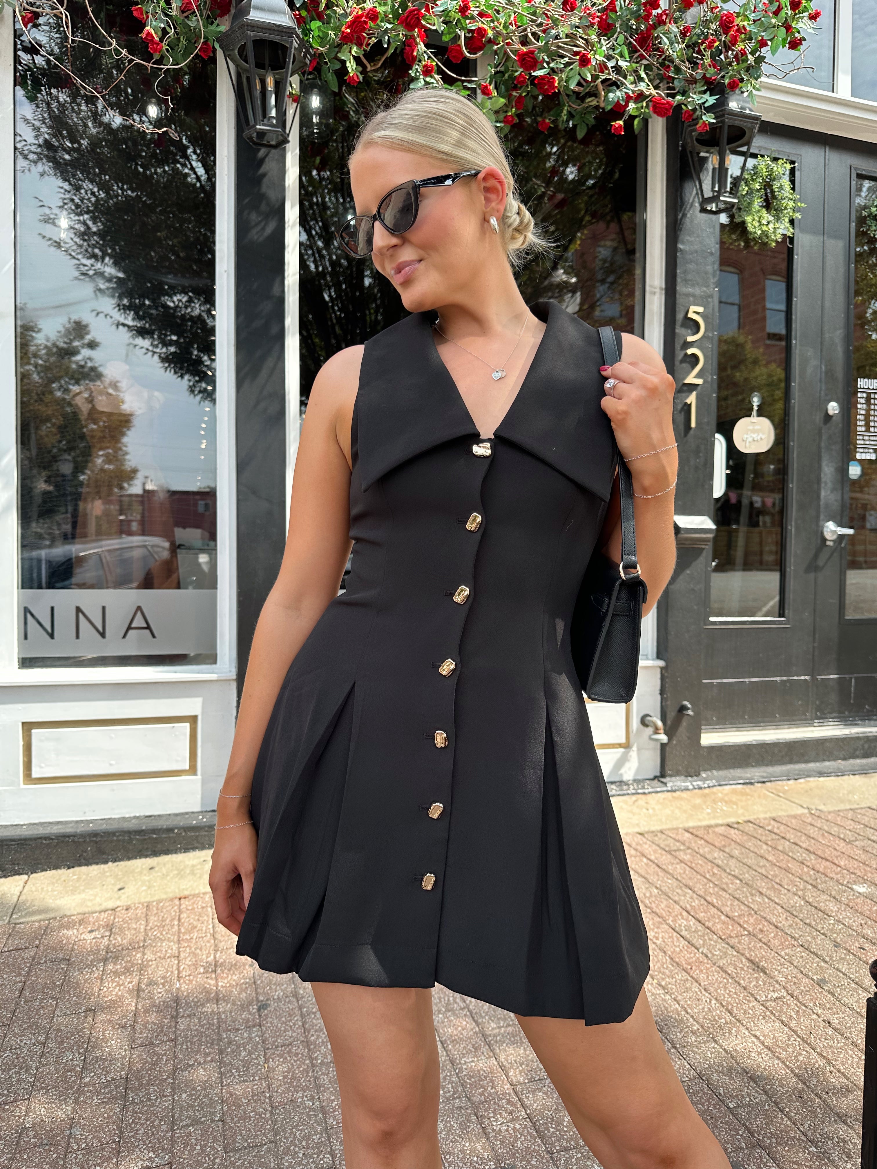 Woman wearing black tailored mini dress with oversized collar neckline and gold button closure. 