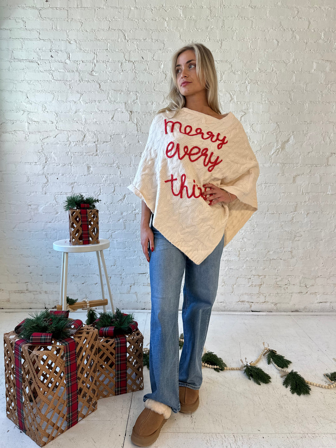 Woman wearing an ivory cable knit poncho with the words merry everything in red cursive. 