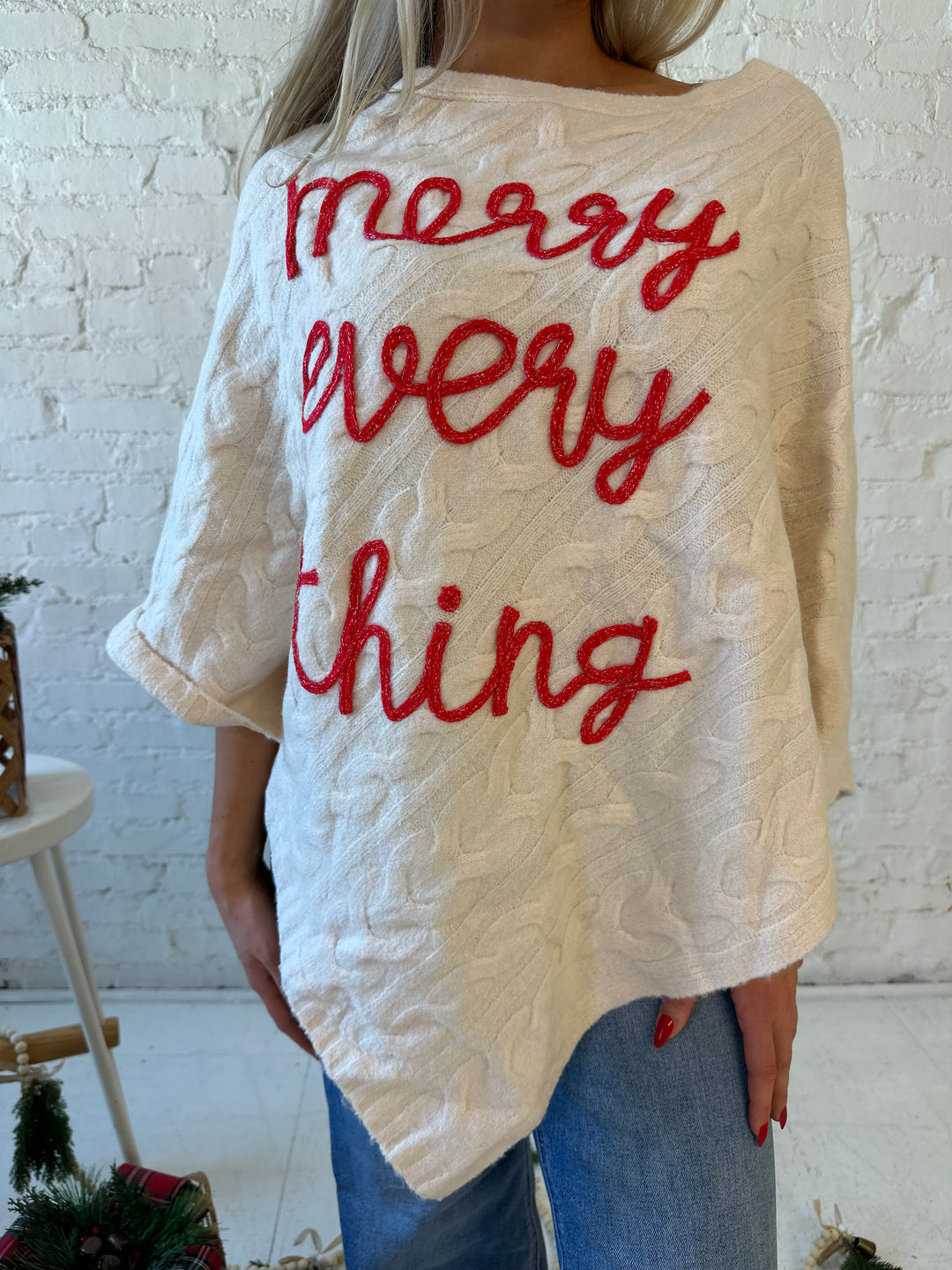 Woman wearing an ivory cable knit poncho with the words merry everything in red cursive. 