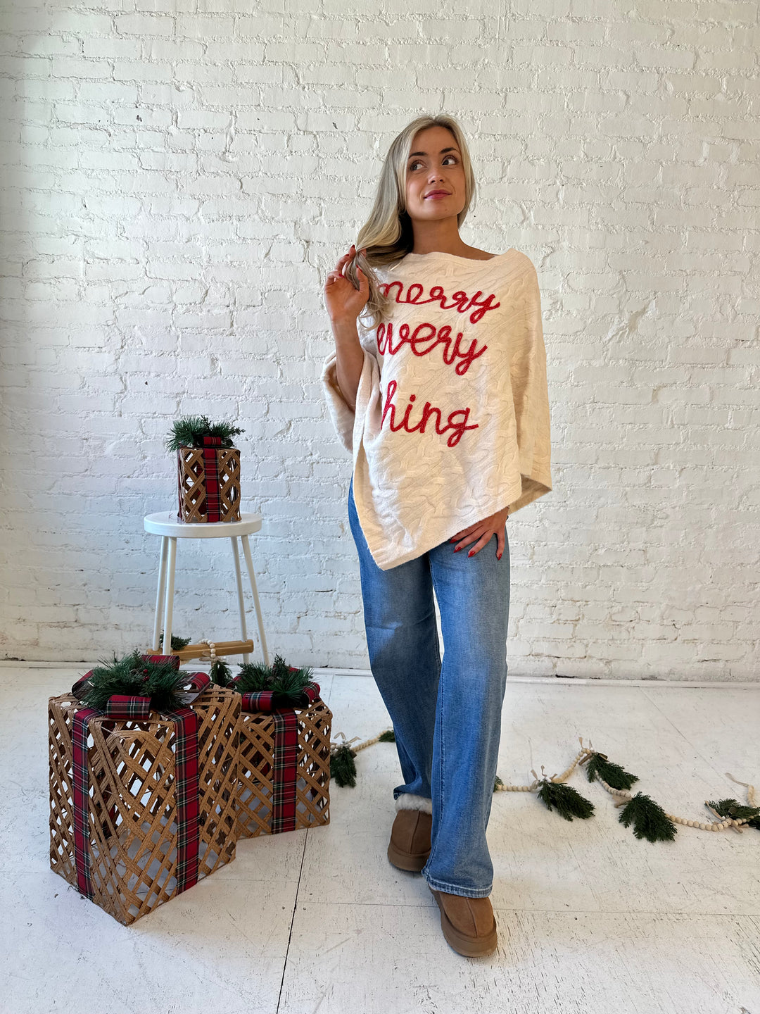 Woman wearing an ivory cable knit poncho with the words merry everything in red cursive. 