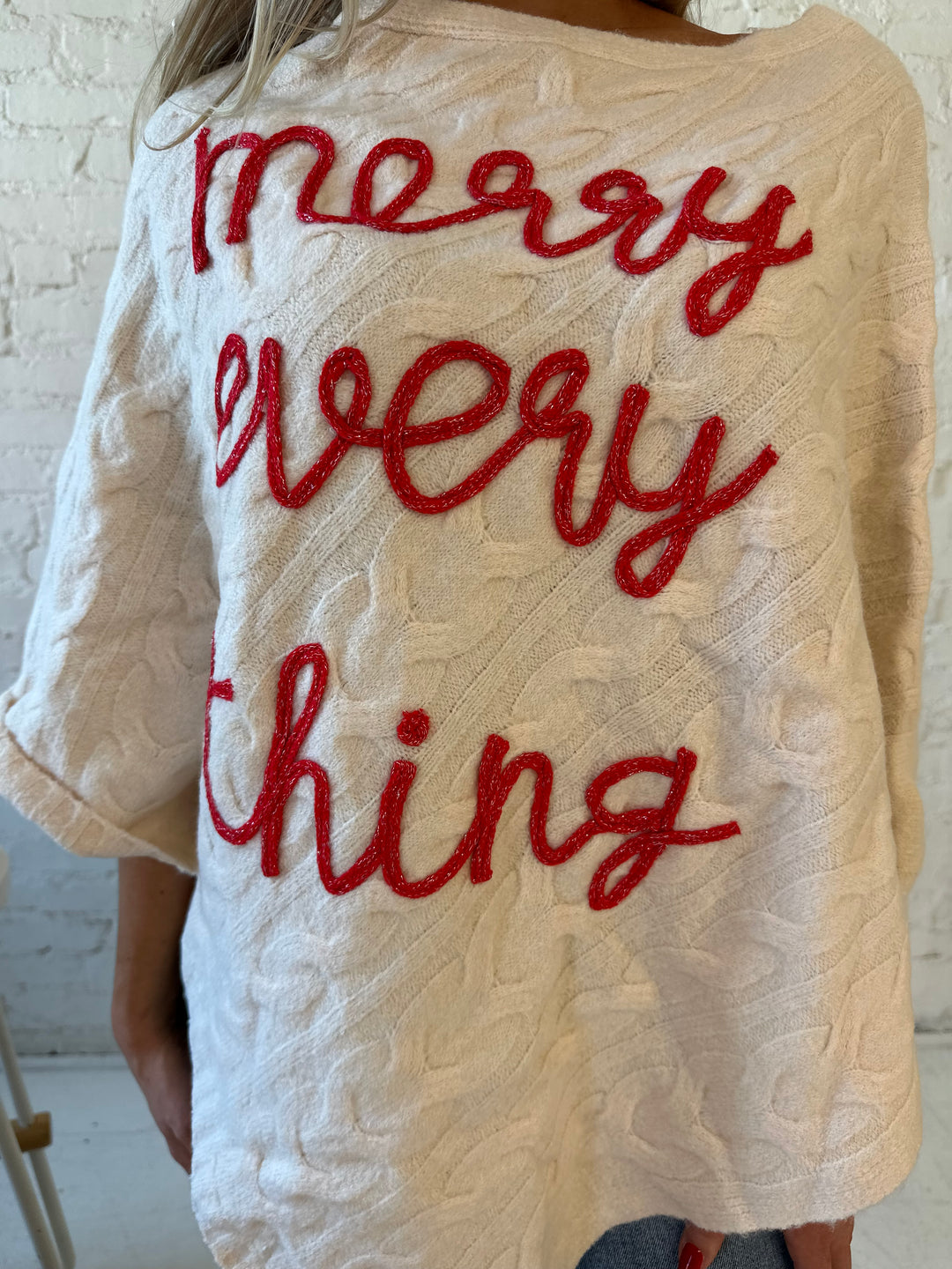 Woman wearing an ivory cable knit poncho with the words merry everything in red cursive. 